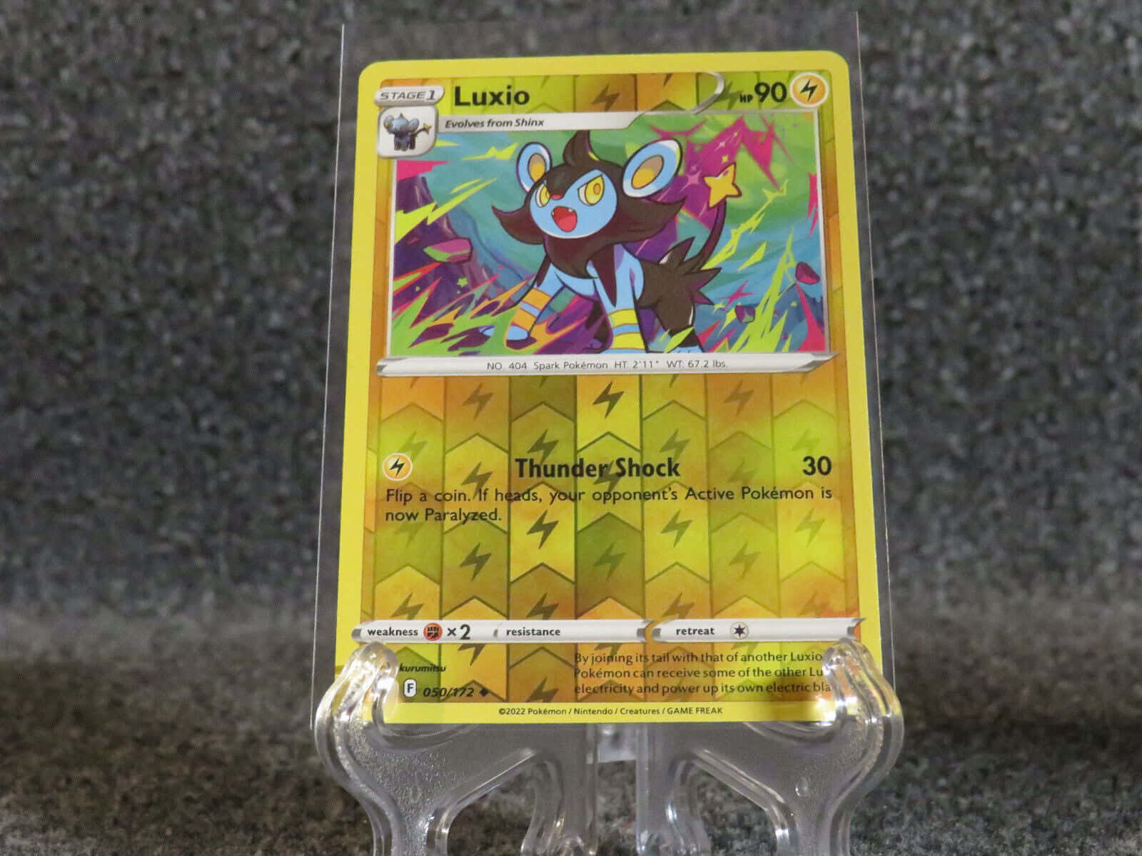 Luxio 寶可夢卡牌展示於透明支架 壁紙 — 一張鮮黃色的Luxio寶可夢卡牌放在透明展示架上，背景為灰色紋理。