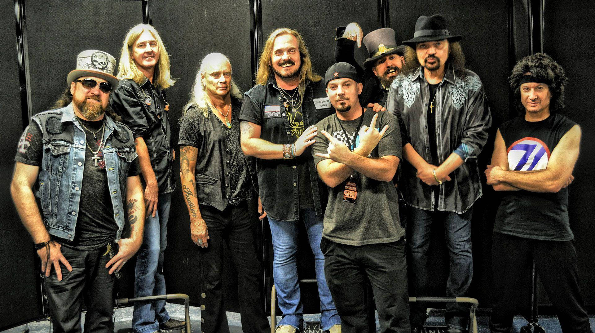 美國搖滾樂團Lynyrd Skynyrd幕後合影 壁紙 — 七位身穿皮背心、戴帽子的搖滾音樂家，在黑色背景前自信地擺姿勢，旁邊有舞台設備。
