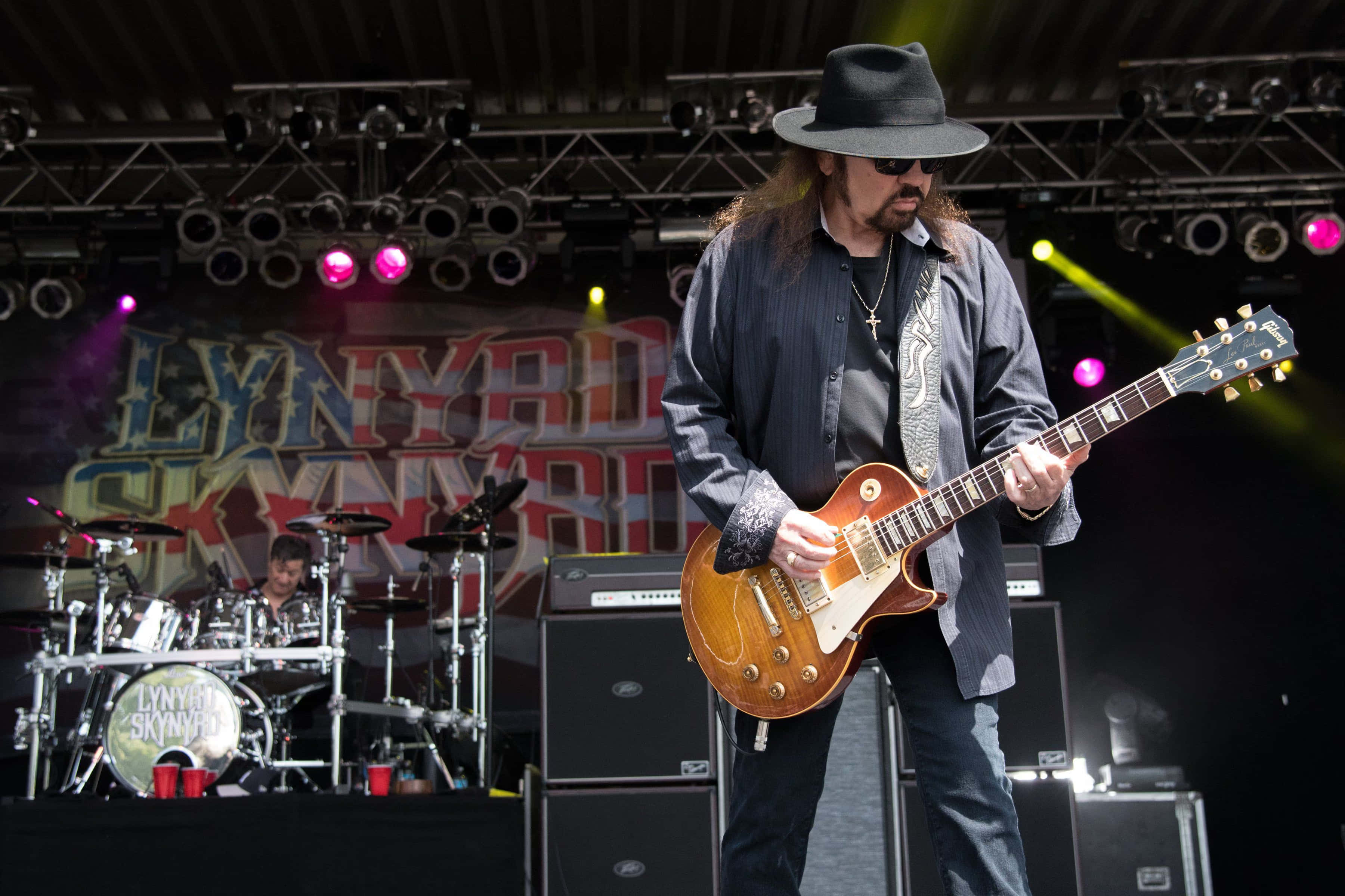 Lynyrd Skynyrd 的 Gary Rossington 壁紙 — 一位戴黑色帽子和太陽眼鏡的結他手在音樂會舞台上彈奏一把 sunburst 電結他，身後有鼓手和色彩繽紛的舞台燈光。