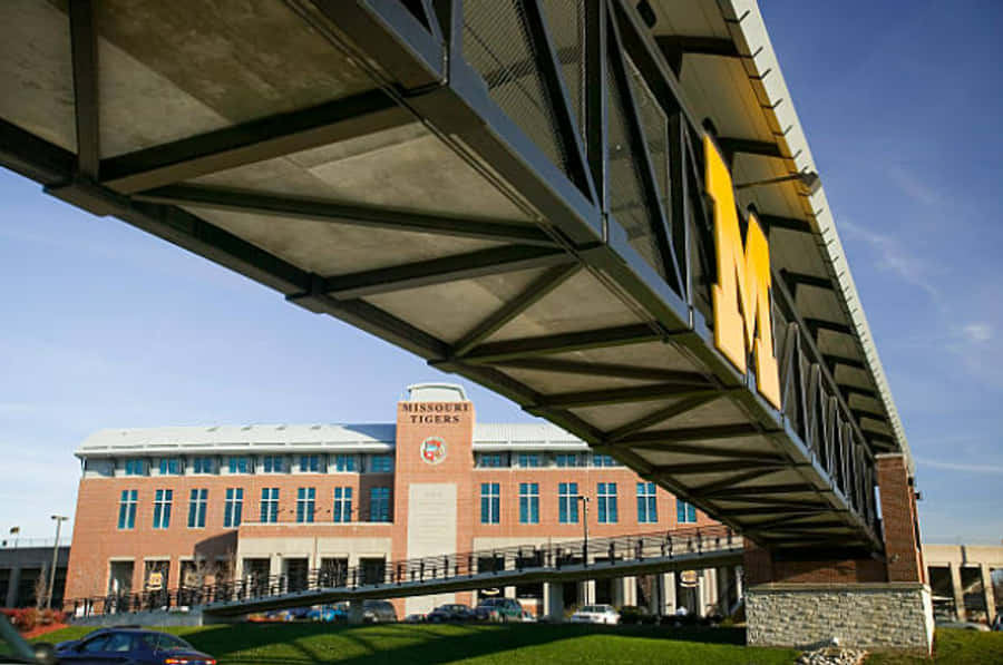 密蘇里大學校園大M字步道 壁紙 — 一個低角度視角，展示一座現代鋼橋，上面有一個巨大的黃色M標誌，橫跨在一棟磚造大學建築上方，背景是清澈的藍天。