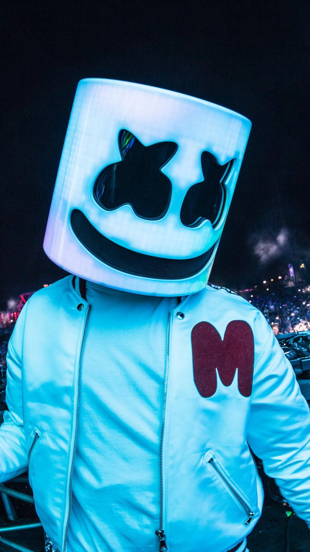 M 字標誌 Bomber Jacket Marshmello iPhone 壁紙 — 一位表演者戴著發光的藍色棉花糖頭盔和帶有紅色M標誌的白色外套，站在黑暗的舞台上，面對人群。