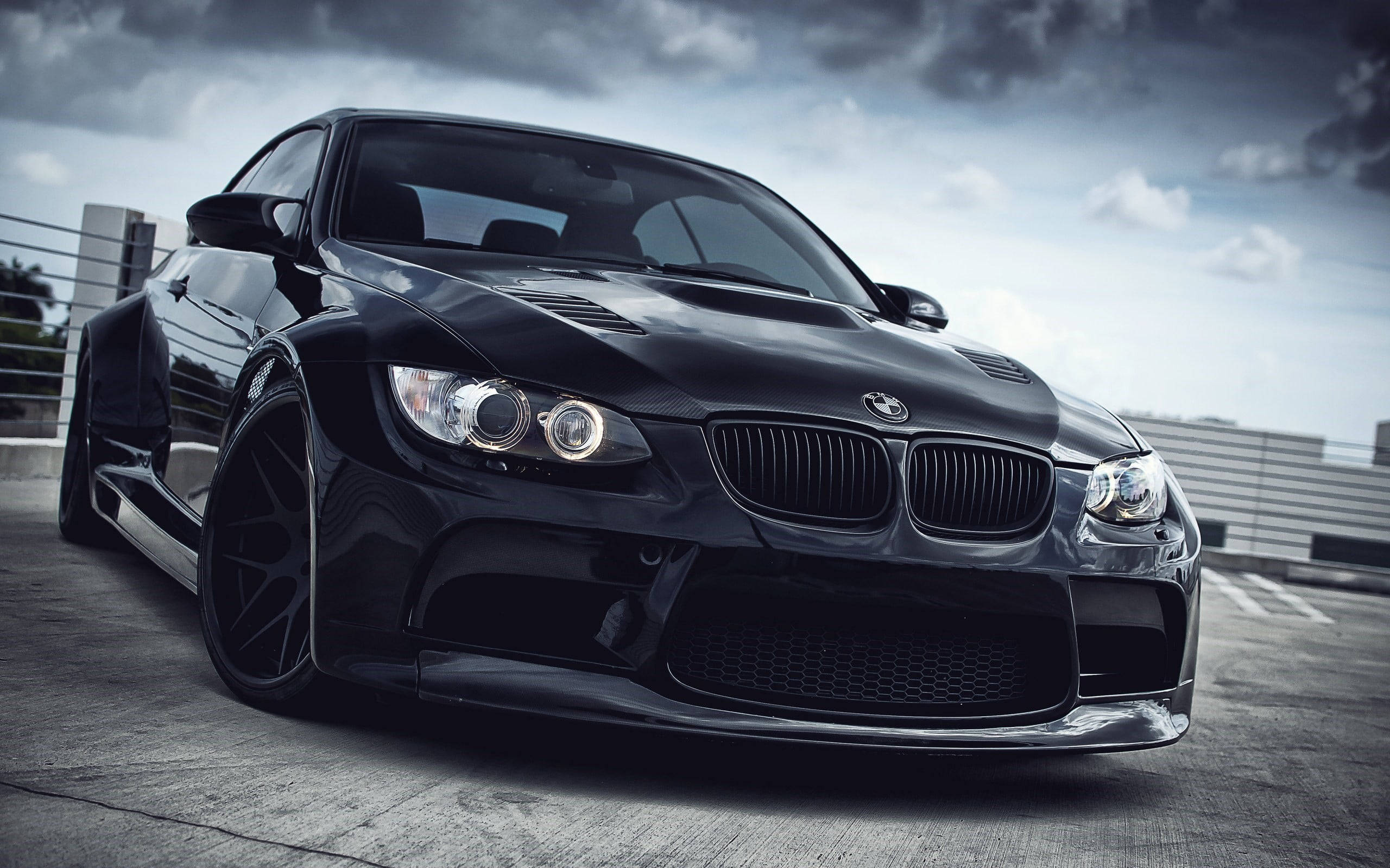 M3轎車BMW筆記本電腦 壁紙 — 一輛時尚的黑色BMW M3跑車，配備定制寬體套件和發光的頭燈，停在混凝土屋頂上，天空多雲而戲劇化。
