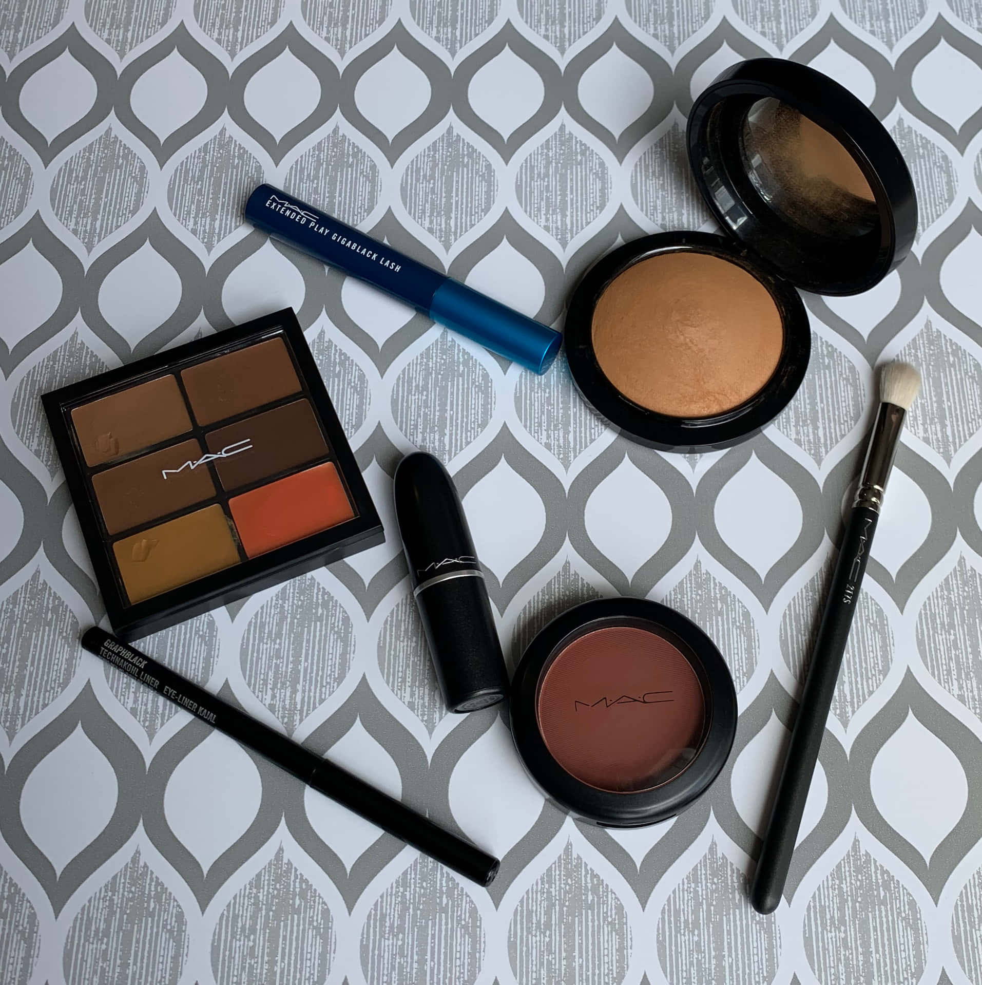 Mac Cosmetics 精緻彩妝陳列 壁紙 — 一個扁平佈局的MAC化妝品，包括眼影、腮紅和唇膏，排列在灰色和白色圖案表面上。