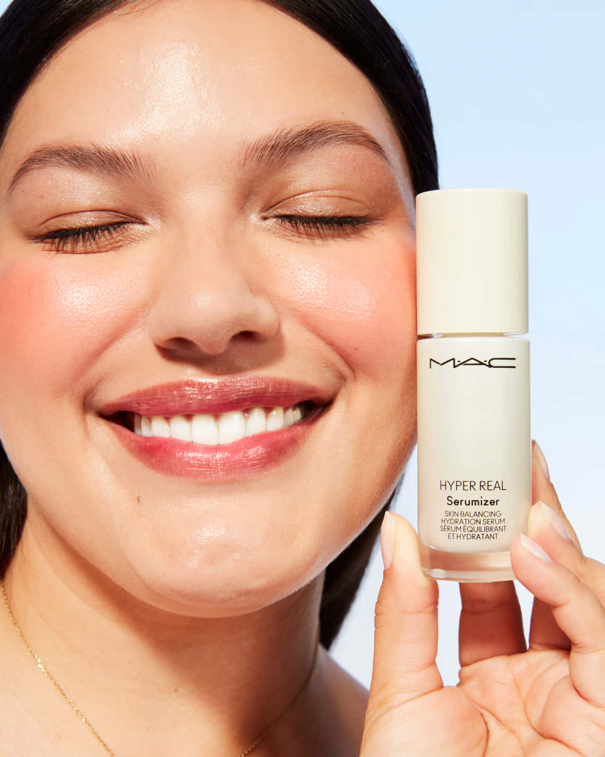 自信出發，盡顯美麗 — Mac Cosmetics 壁紙 — 一位微笑的女子閉著眼睛，手持MAC Hyper Real Serum瓶子，背景是柔和的藍色。