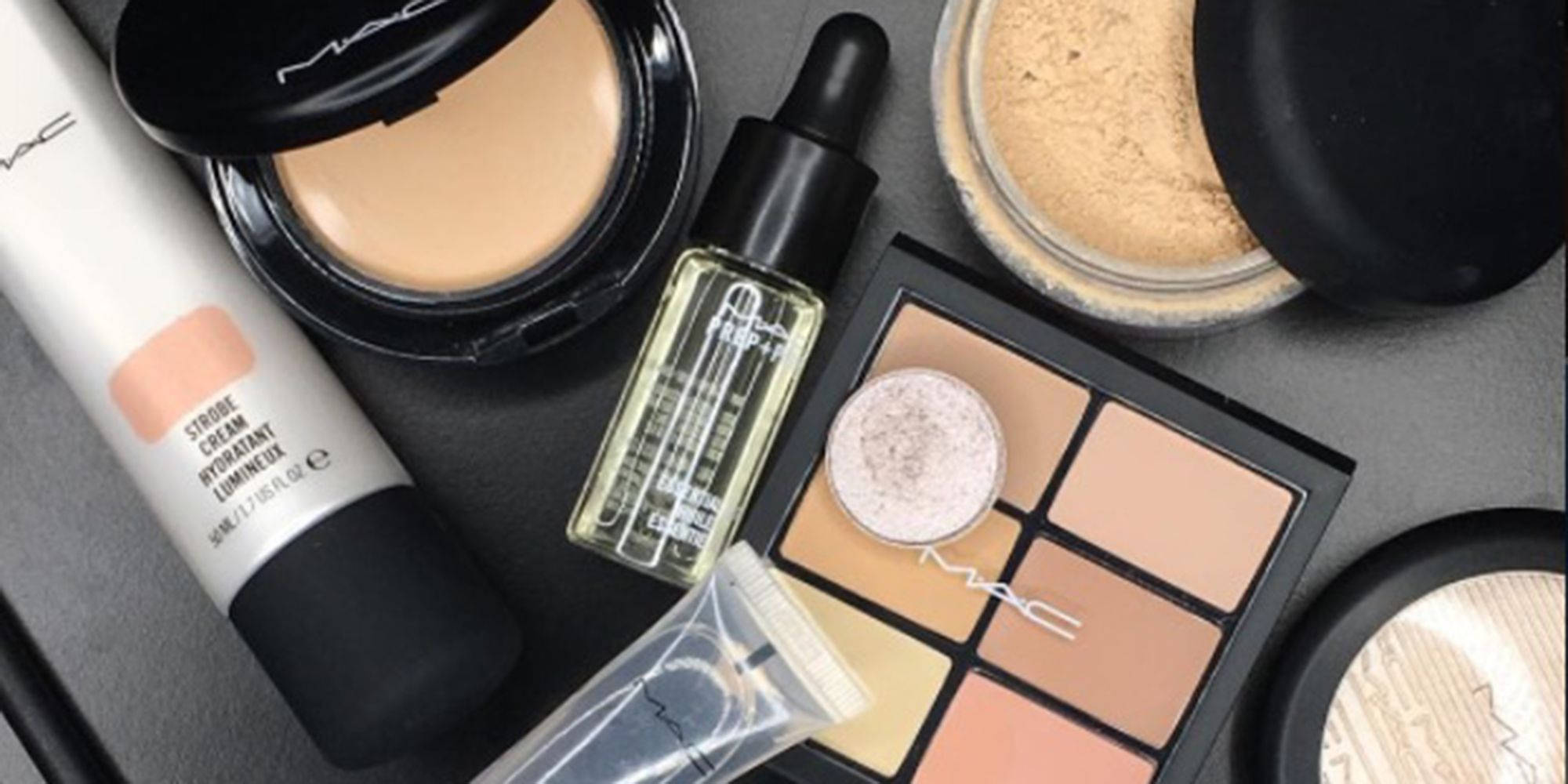 Mac Cosmetics 無瑕光感肌膚 壁紙 — 在深灰色表面上平鋪擺放的MAC化妝品，包括粉底、遮瑕膏、精華液和眼影盤。