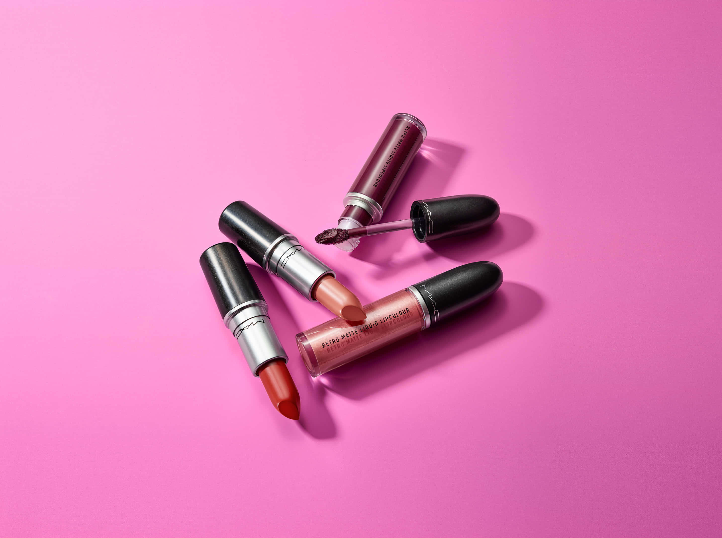 以 MAC 化妝品提升您的美麗 壁紙 — 四款暖色調的MAC唇部產品散落在充滿活力的粉紅色背景上，帶有柔和陰影。