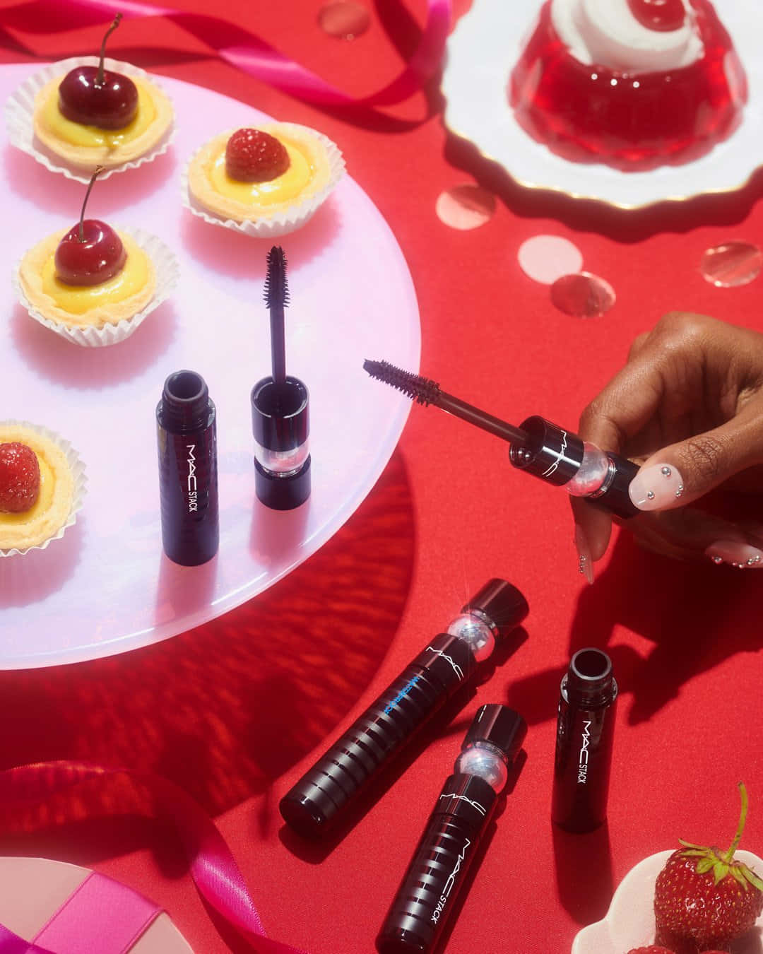 以 MAC 化妝品展現你的美麗 壁紙 — 一隻帶有閃亮指甲的手在一個散落著迷你撻、櫻桃和果凍甜點的鮮紅色表面上拿著一支MAC睫毛膏棒。