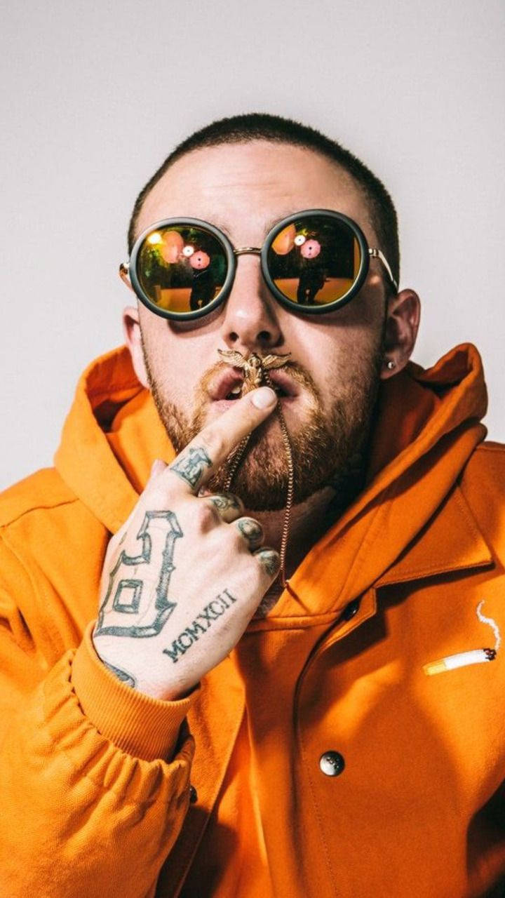 Mac Miller 戴項鍊於嘴 壁紙 — 一名身穿亮橙色連帽衫和圓形太陽眼鏡的男子，將香煙放在唇邊，直視鏡頭。