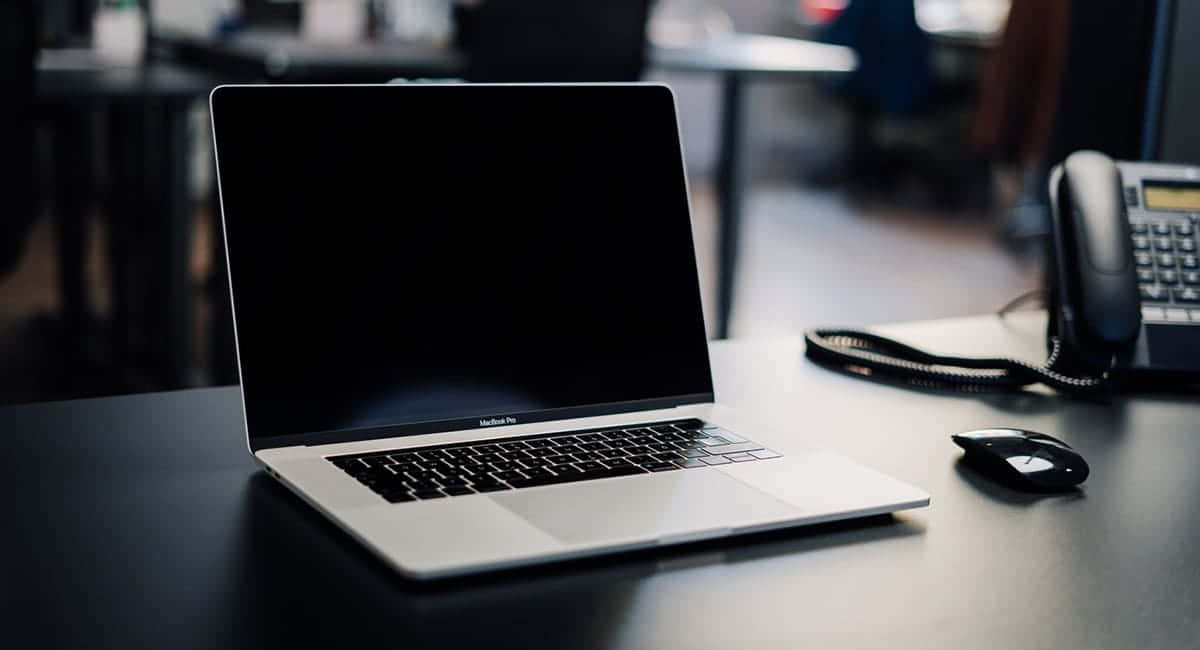 專為創作者打造的高階黑色MacBook 壁紙 — 一部時尚的銀色 MacBook Pro 打開放在深色桌子上，旁邊有一個黑色滑鼠和一個座機電話，背景是柔和模糊的辦公室環境。