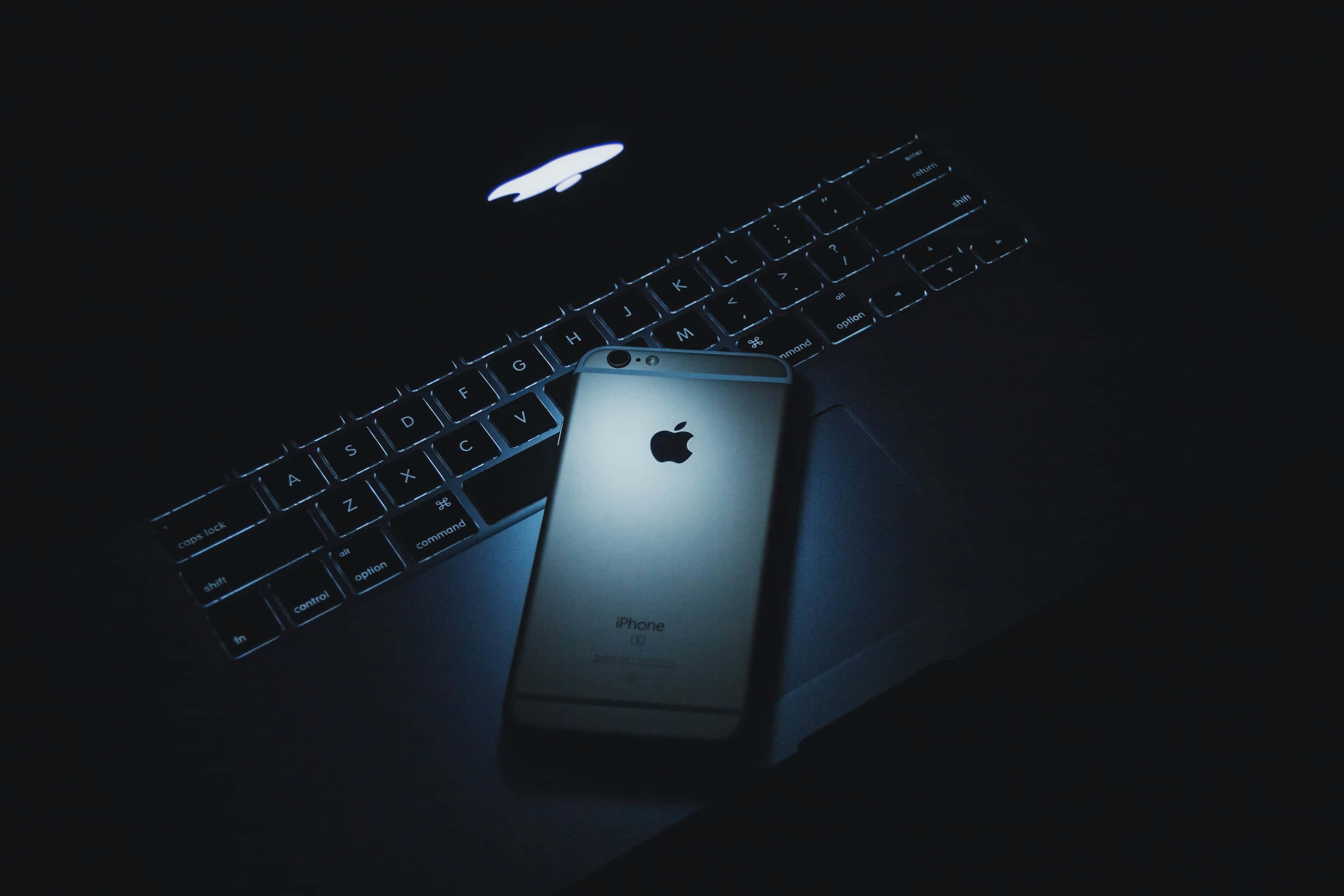 享受舒適的 MacBook 使用體驗 壁紙 — 一部銀色iPhone在冷藍色燈光下放在深色筆記型電腦鍵盤上，Apple標誌發光。