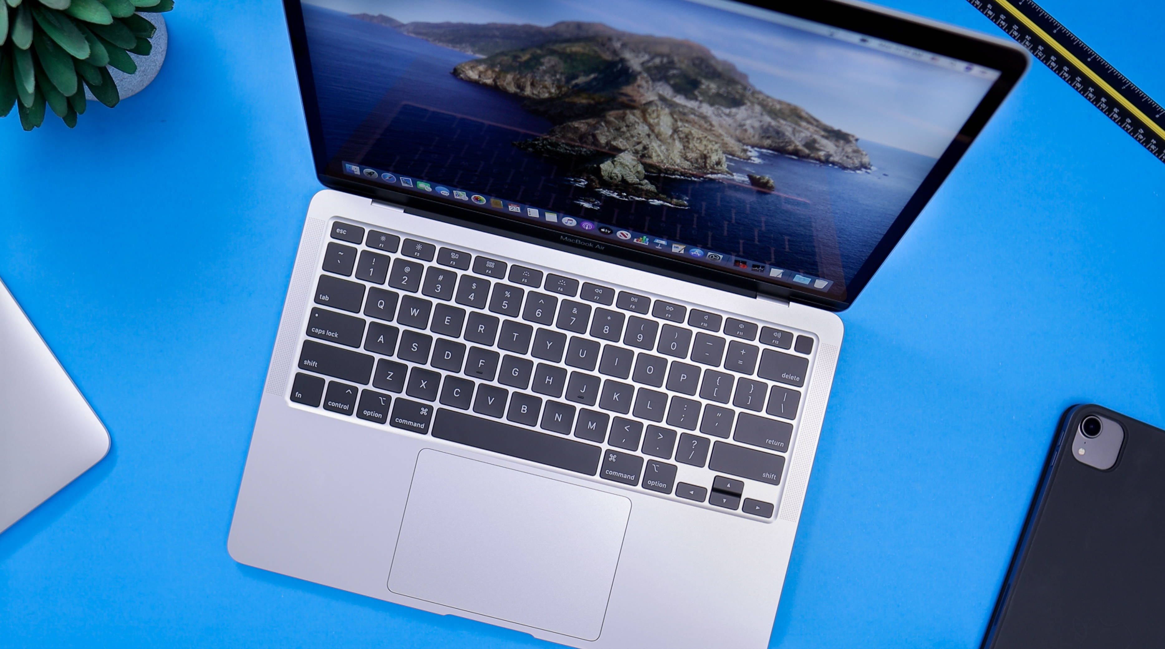 MacBook Air 2020藍色表面 壁紙 — 銀色手提電腦放在亮藍色書桌上，附近有島嶼牆紙和多肉植物。