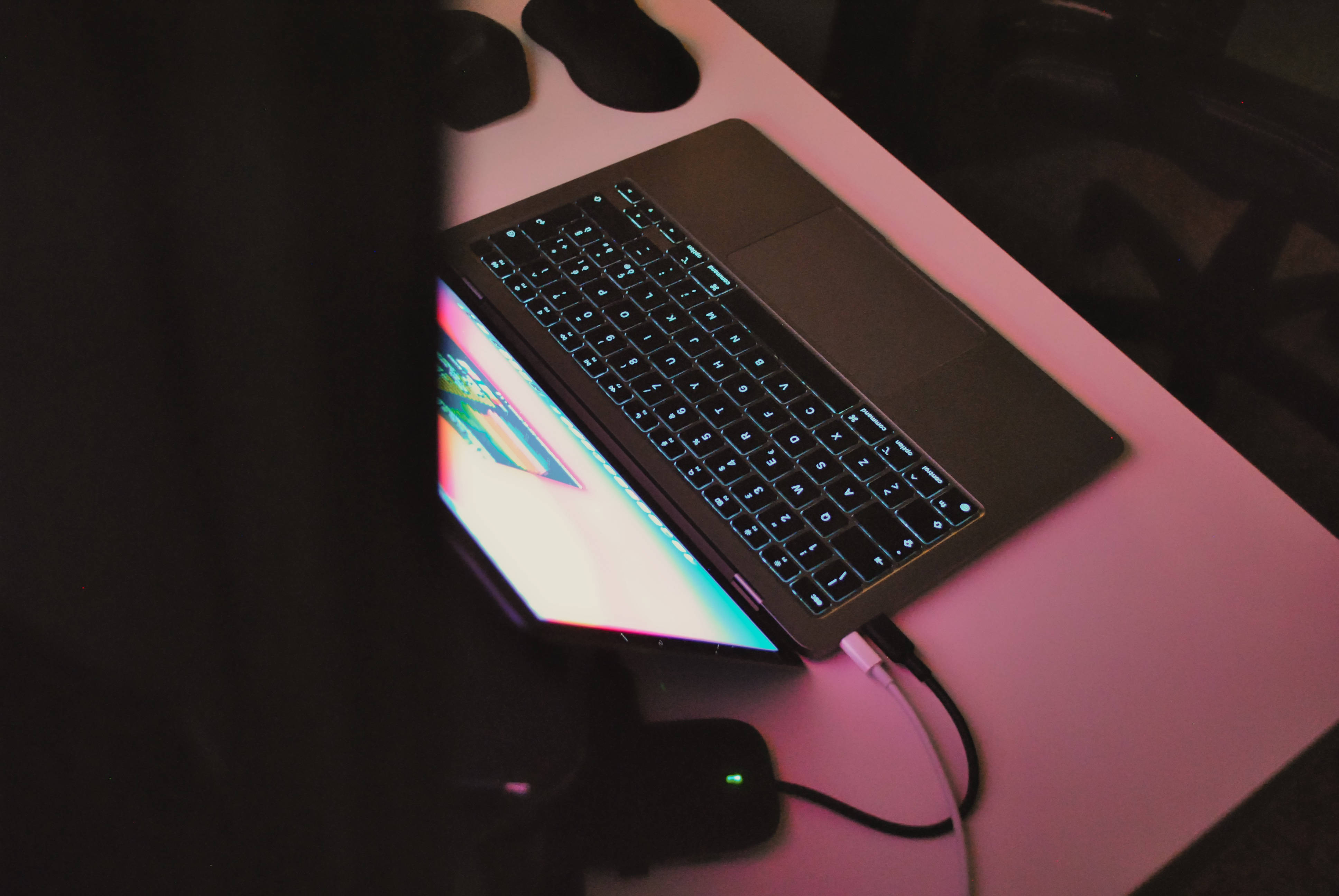 MacBook Air 2020粉紅背光 壁紙 — 一部帶有發光鍵盤的筆記型電腦放在一張被粉紅色燈光籠罩的桌子上，附近有色彩繽紛的螢幕和週邊設備。