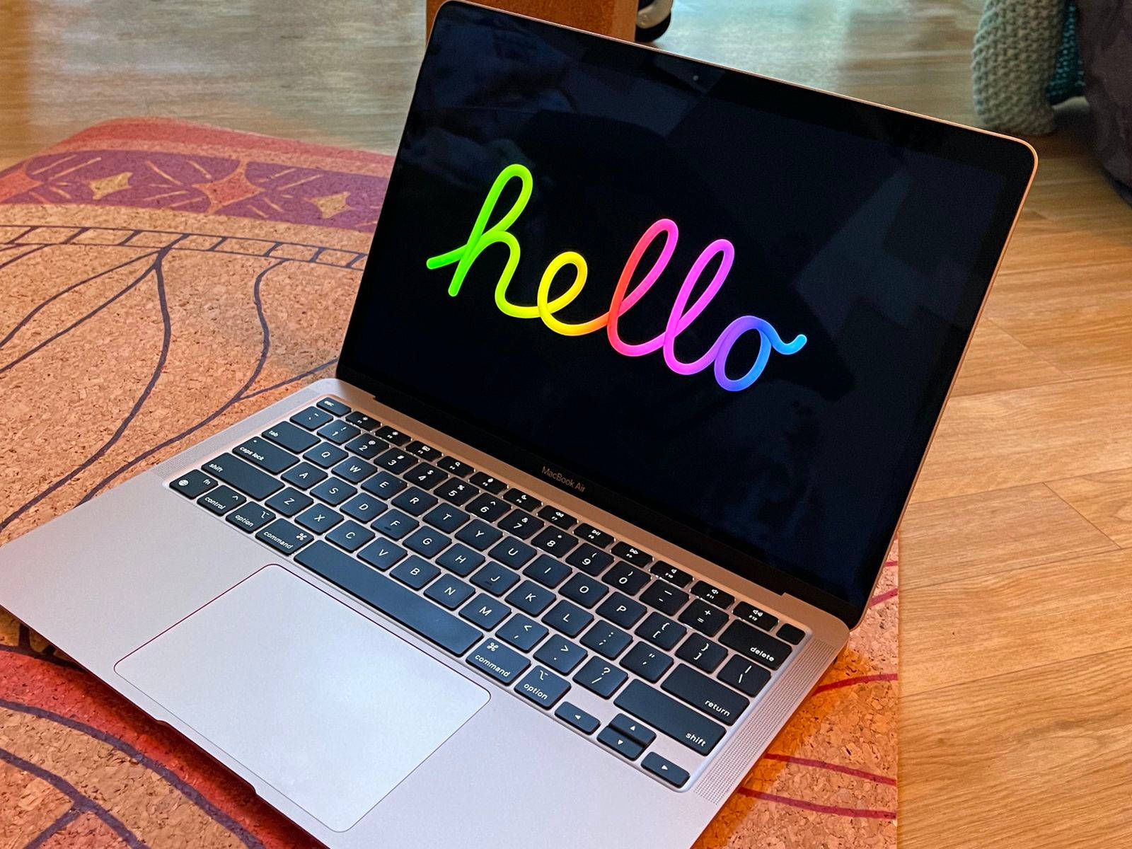 MacBook Air 開機問候畫面 壁紙 — 一部銀色 MacBook Air 在木地板上嘅圖案地毯上，螢幕顯示彩虹漸變嘅哈囉。