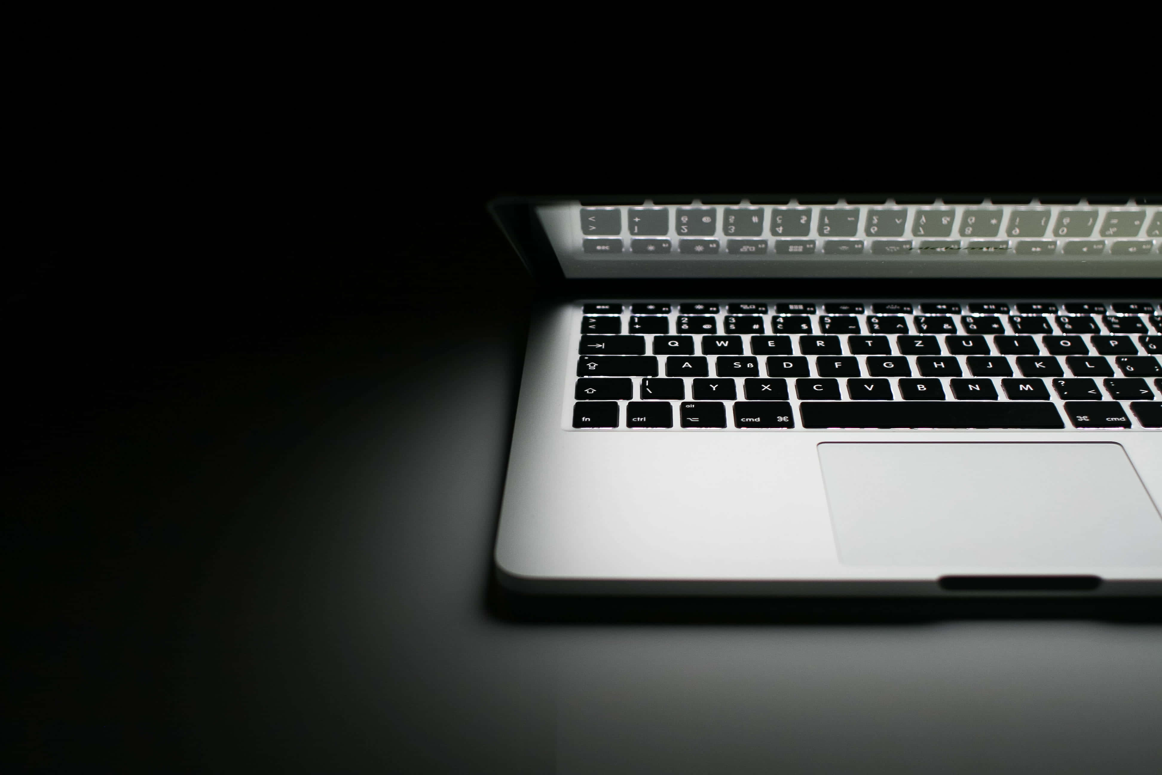 美學 MacBook - 工作時展現風格 壁紙 — 時尚的銀色筆記型電腦鍵盤在深黑色背景上柔和發光，帶有戲劇性的聚光燈效果。