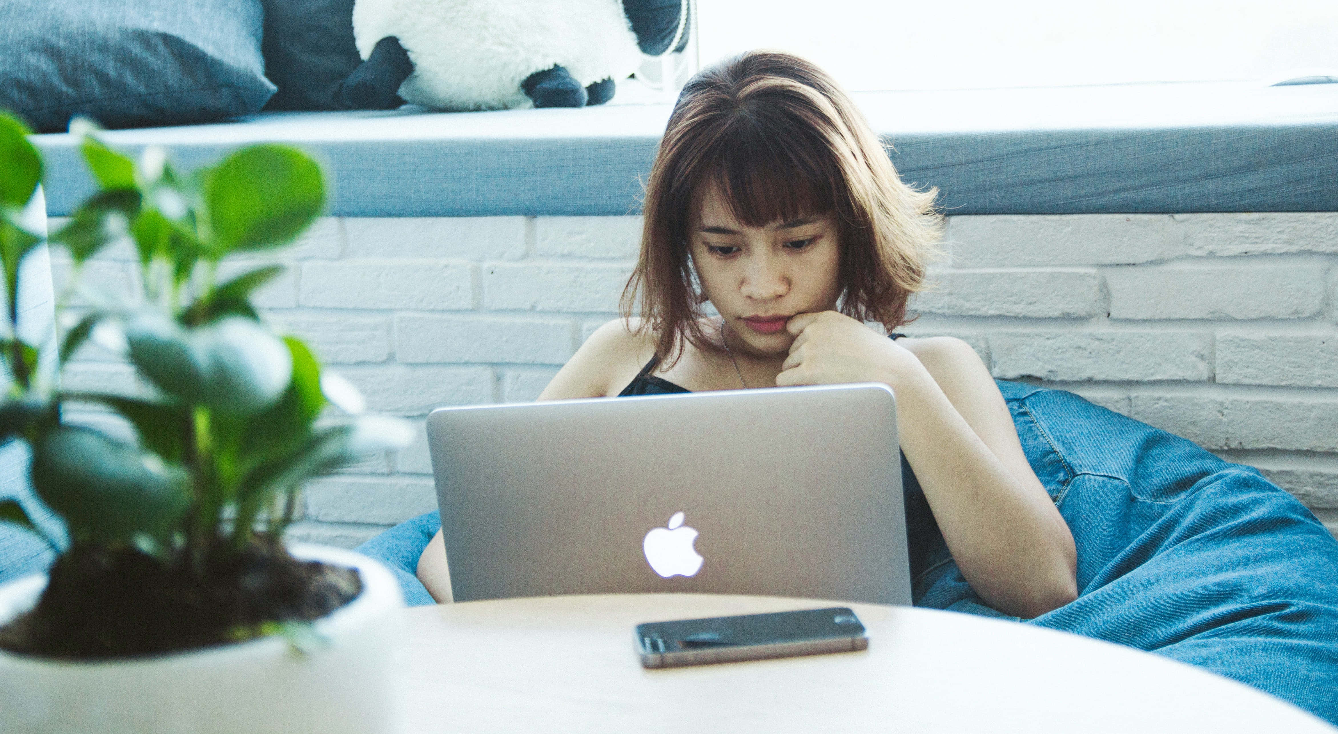 明亮靈感粉大理石美學MacBook 壁紙 — 一位棕色頭髮的年輕女性坐在藍色豆袋椅上，專注於她的筆記型電腦，房間明亮，附近有一盆盆栽植物。