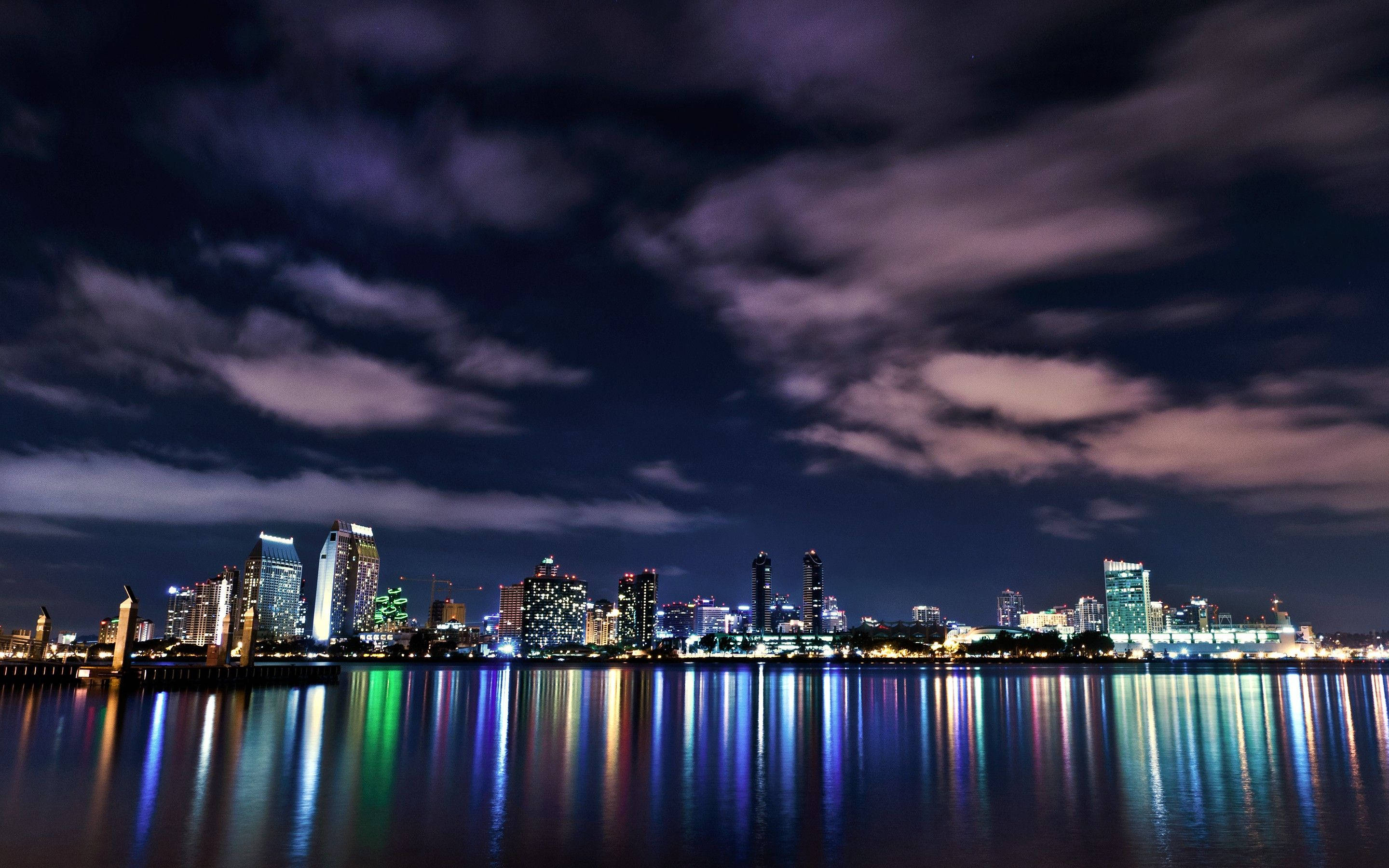MacBook Pro Glowing Cityscape 壁紙 — 在戲劇性的多雲天空下，充滿活力的城市天際線在平靜的水面上倒映。