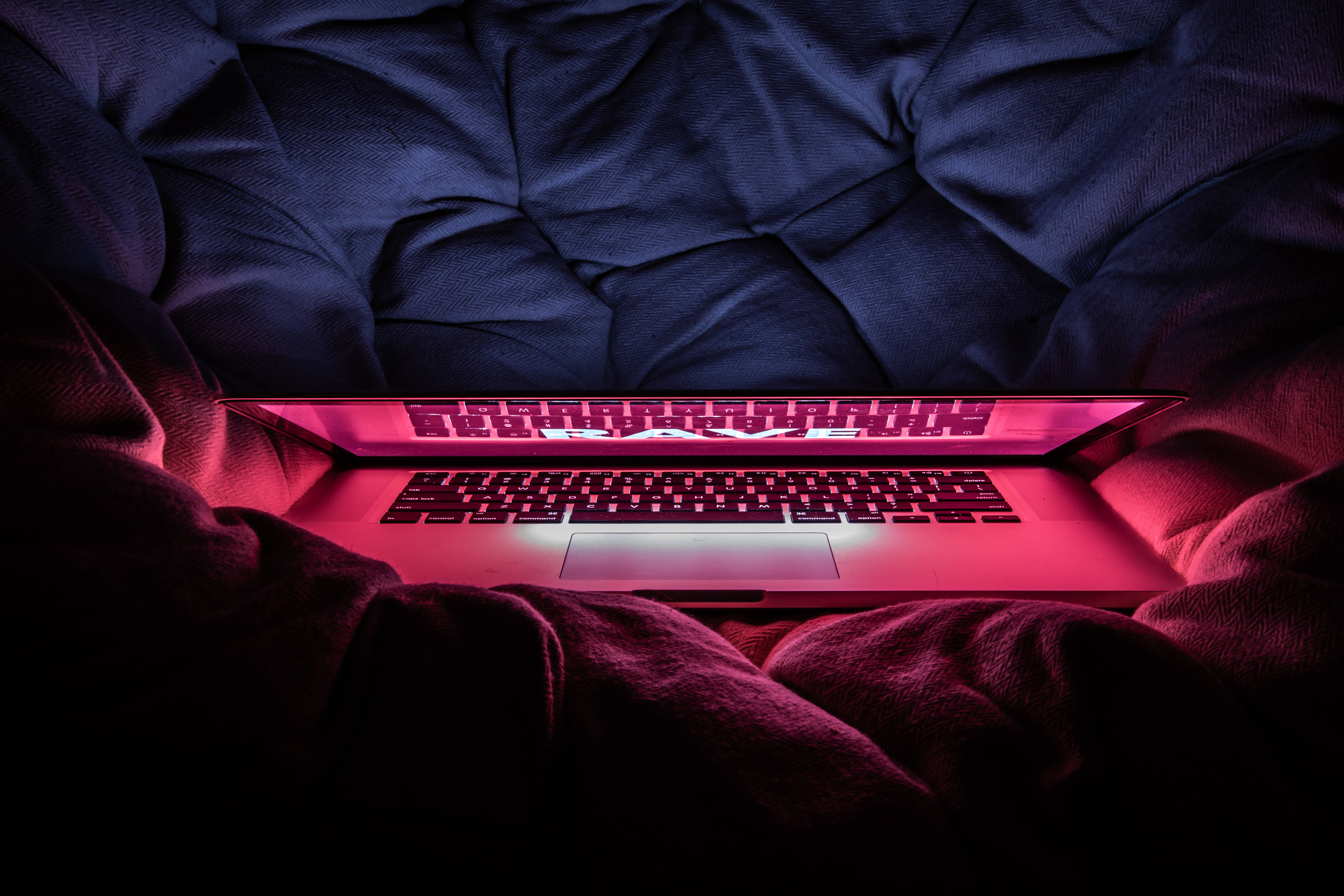 MacBook Pro 紅色派對 壁紙 — 一部發出粉紅色光的筆記型電腦放在昏暗房間中皺巴巴的深藍色床單上。