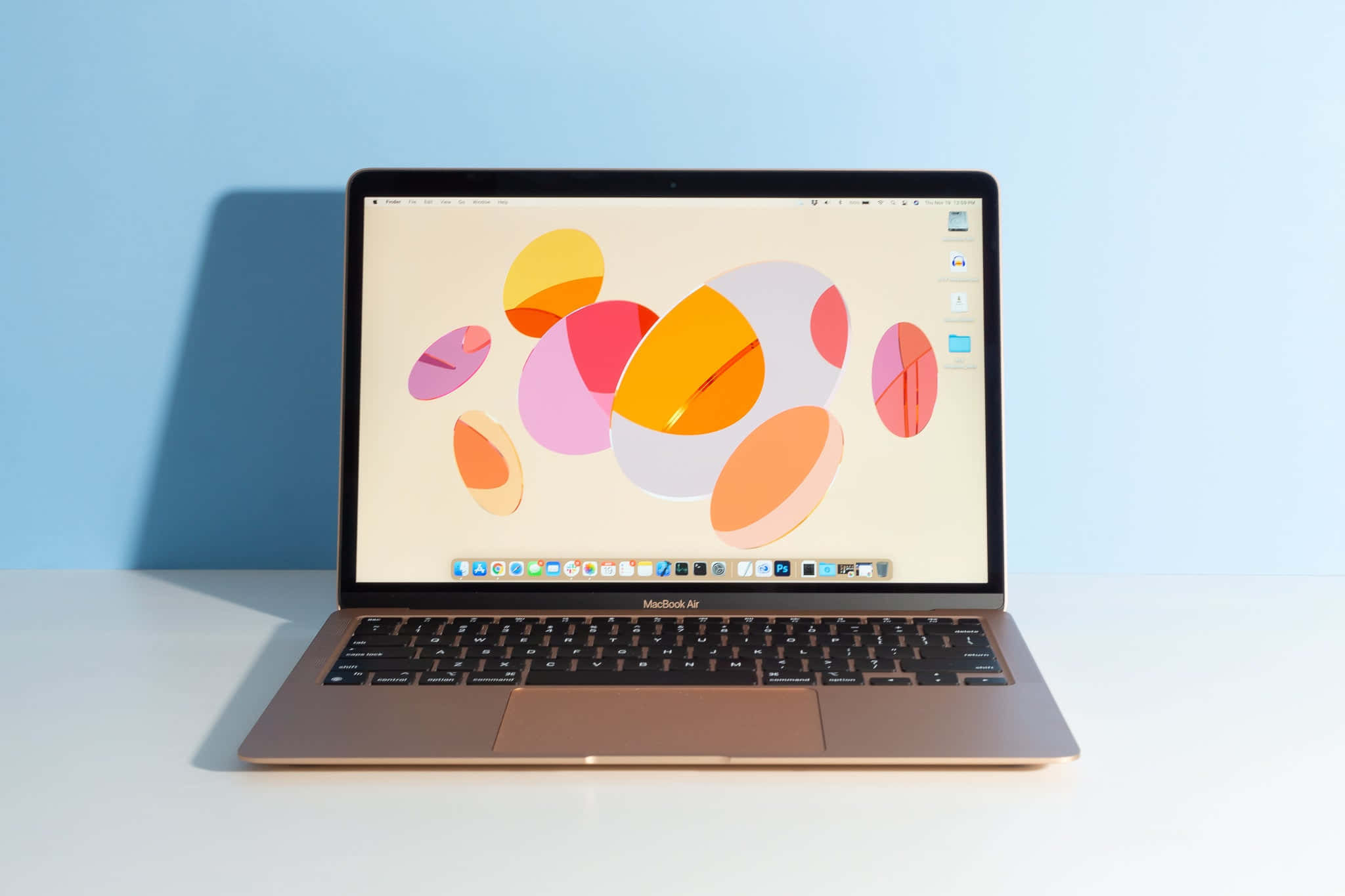 以 MacBook 將生產力提升至新境界 壁紙 — 一部纖薄的金色 MacBook Air 在白色表面上打開，對著柔和的藍色牆壁，顯示充滿活力的抽象桌布，上面有重疊的彩色圓圈。