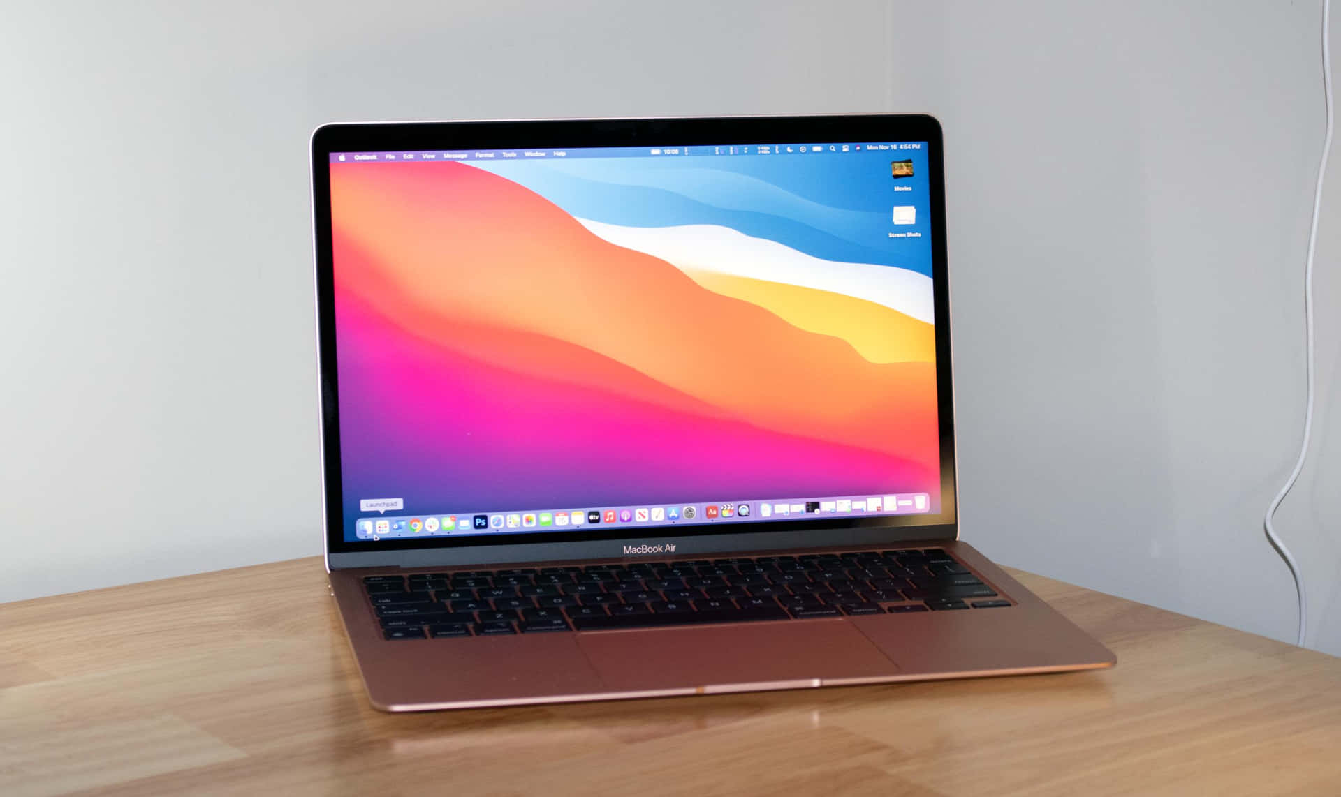 Macbook 前方擺放，透過鋁合金機身欣賞城市風景 壁紙 — 一部玫瑰金色 MacBook Air 打開放在木桌上，顯示一幅充滿活力的抽象桌布，帶有粉紅、橙色和藍色的流動漸變。