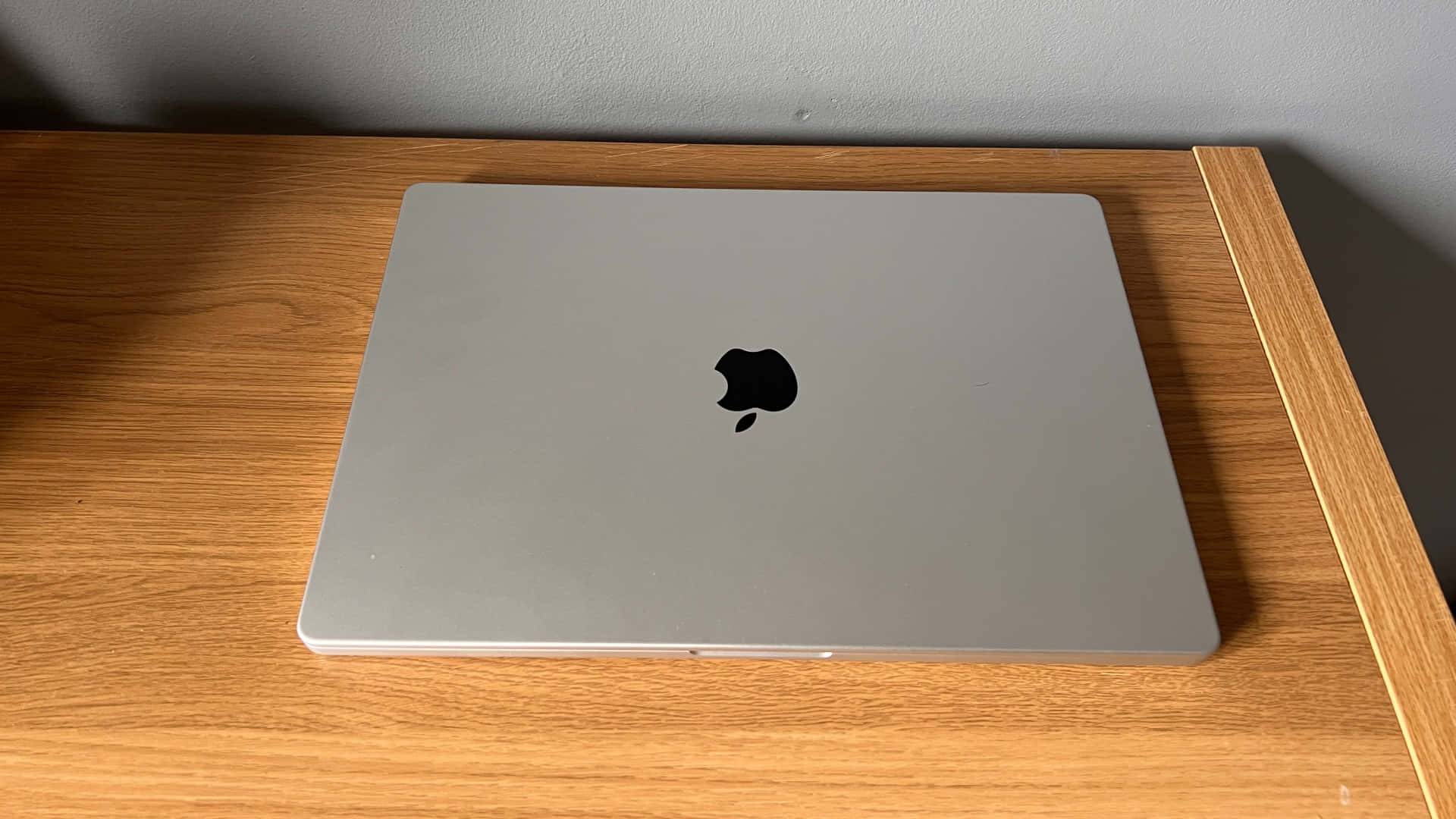 MacBook —— 開啟數位世界的門戶 壁紙 — 一部關閉的銀色MacBook放在淺色木桌上，背景是純灰色牆壁。