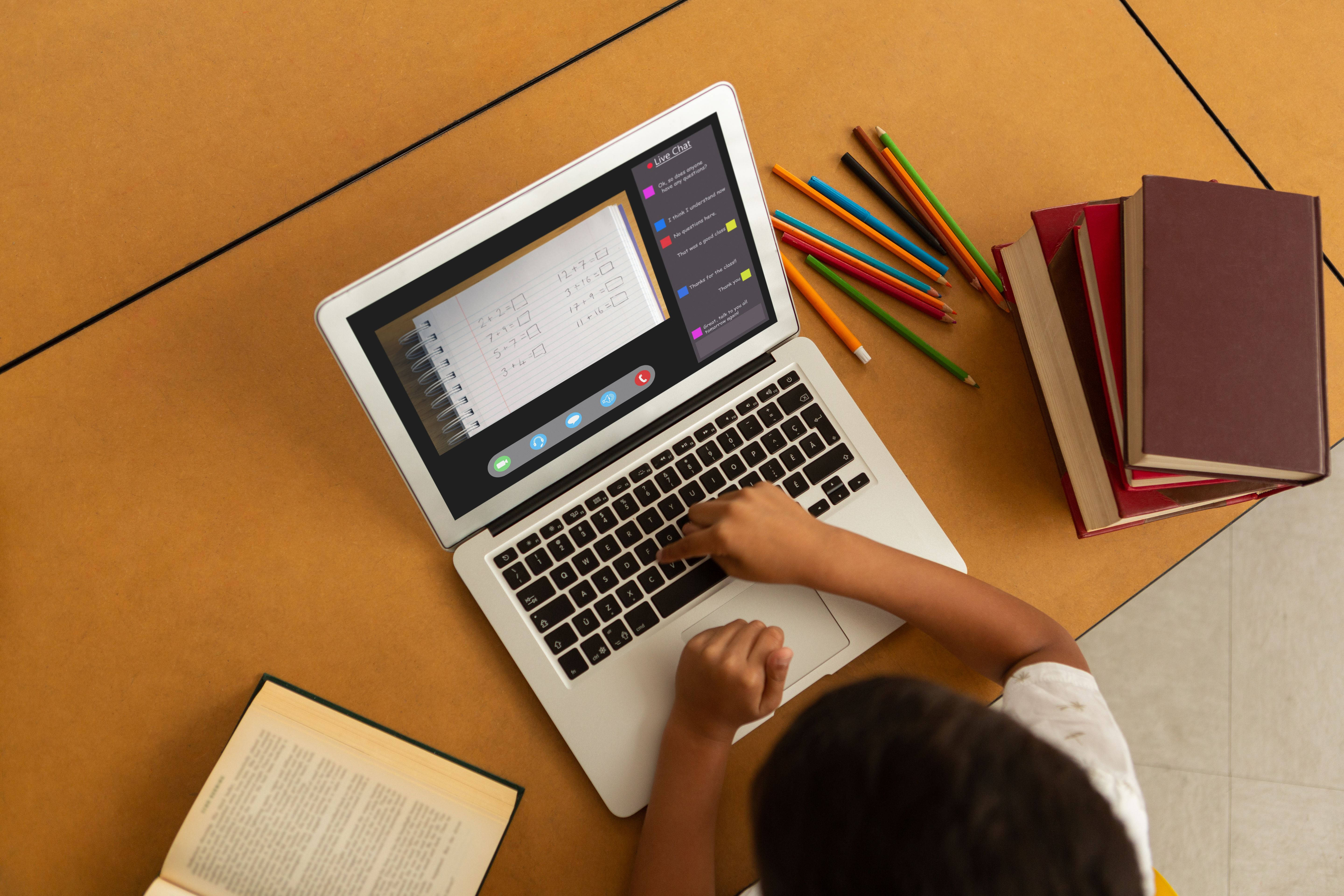 MacBook學習環境 壁紙 — 從上方俯瞰兒童雙手在木桌上打字，周圍環繞著書籍和彩色鉛筆的筆記型電腦。