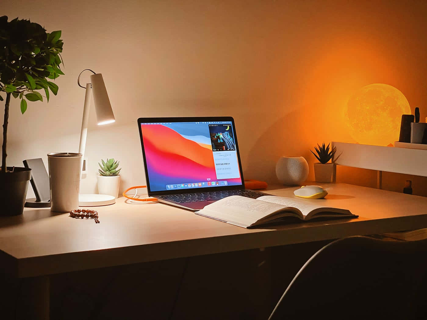 MacBook歡迎您進入創意世界 壁紙 — 一個舒適的辦公桌設置，沐浴在發光的月亮燈和檯燈的溫暖環境光中，有一本打開的書和顯示彩色壁紙的筆記本電腦。