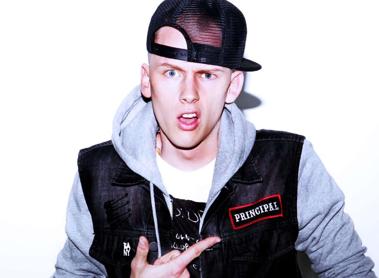 Machine Gun Kelly — 饒舌藝術家 壁紙 — 一名年輕男子戴著黑色網帽和牛仔背心，指著一個「PRINCIPAL」補丁，在白色背景前露出驚訝的表情。