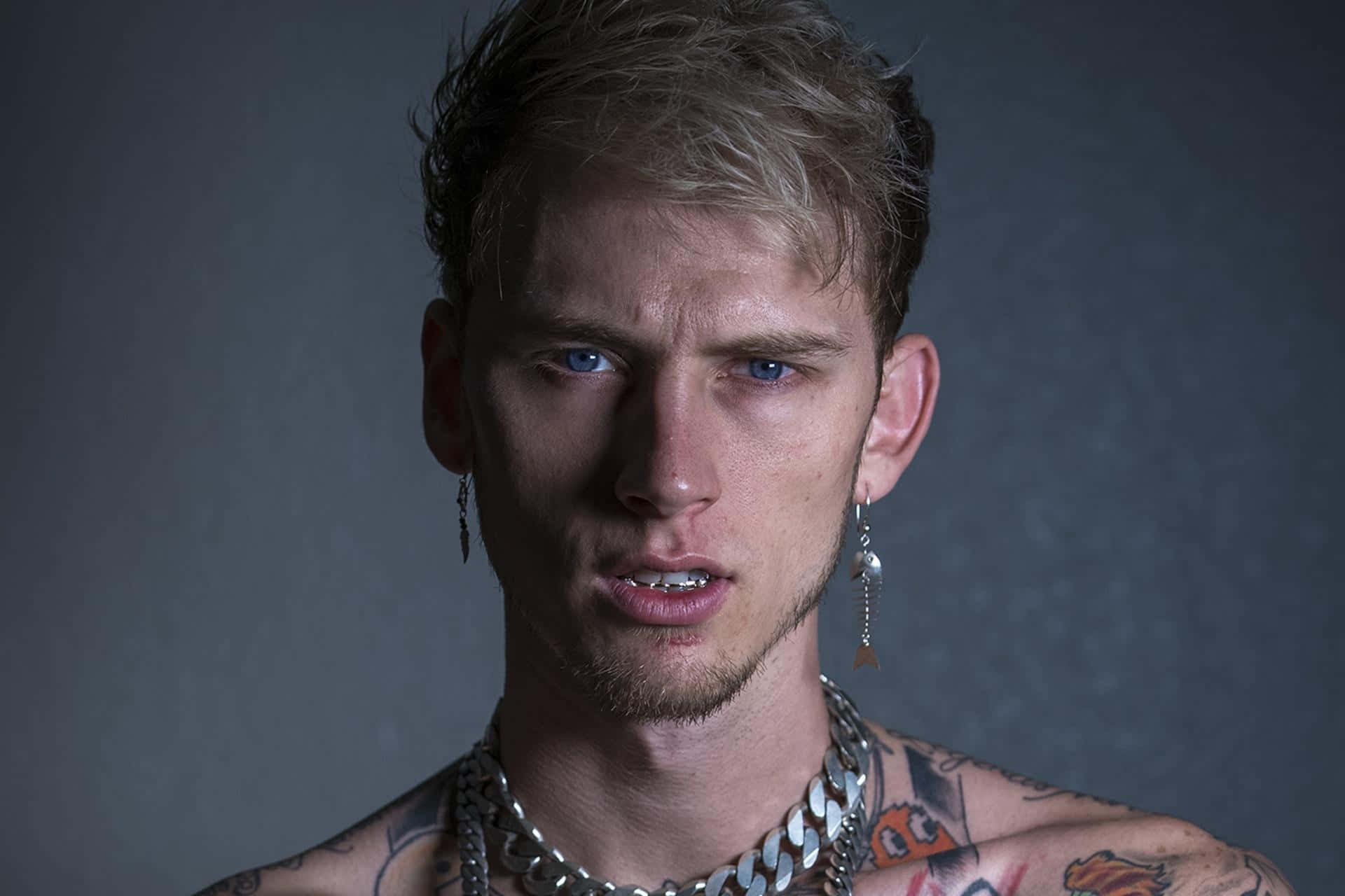 饒舌歌手 Machine Gun Kelly 壁紙 — 一位藍眼睛、有紋身的年輕男子在深灰色背景下強烈凝視。