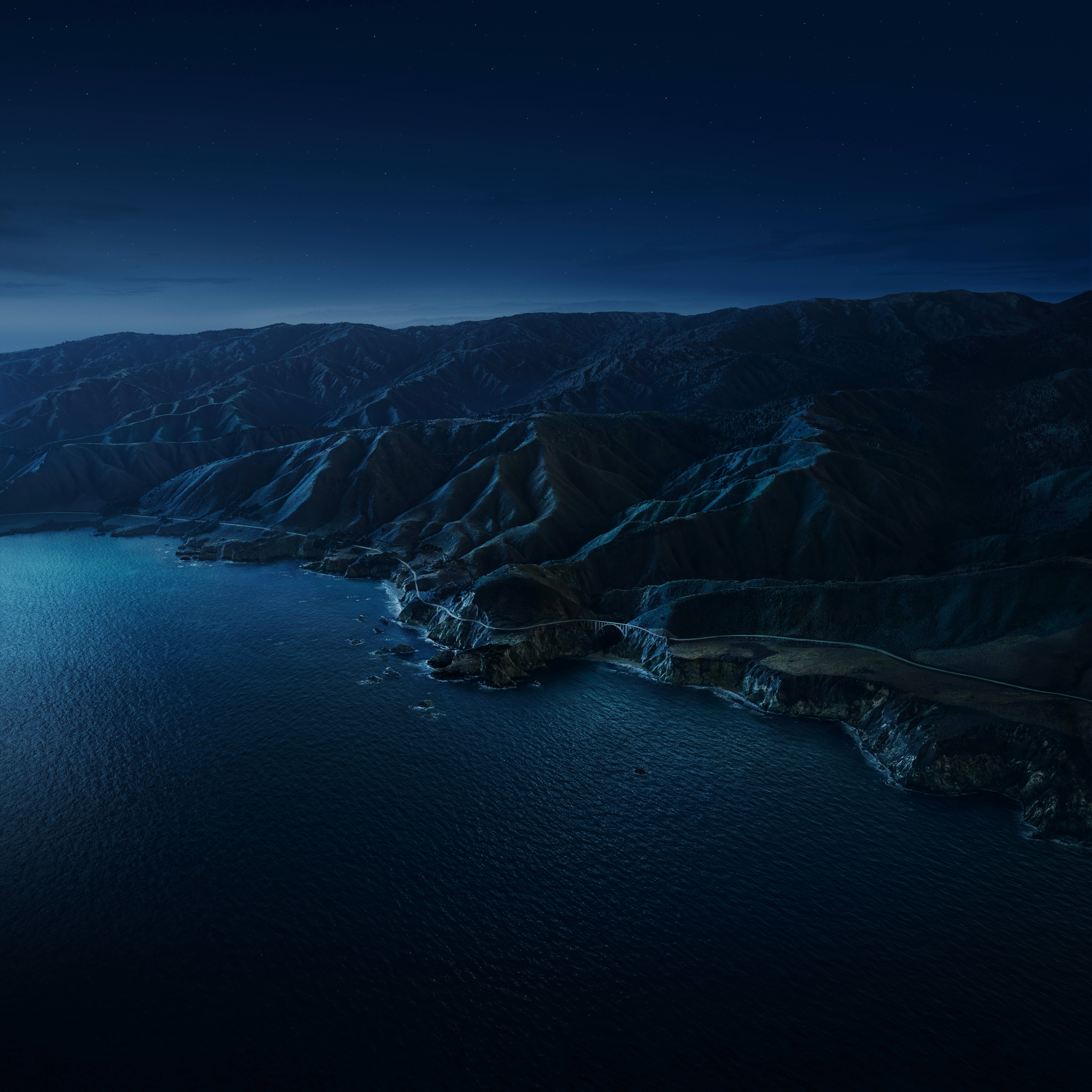 macOS Big Sur 夜間海景 壁紙 — 蜿蜒的海岸公路在深藍色海洋上方的星空夜空下緊貼著黑暗的懸崖。