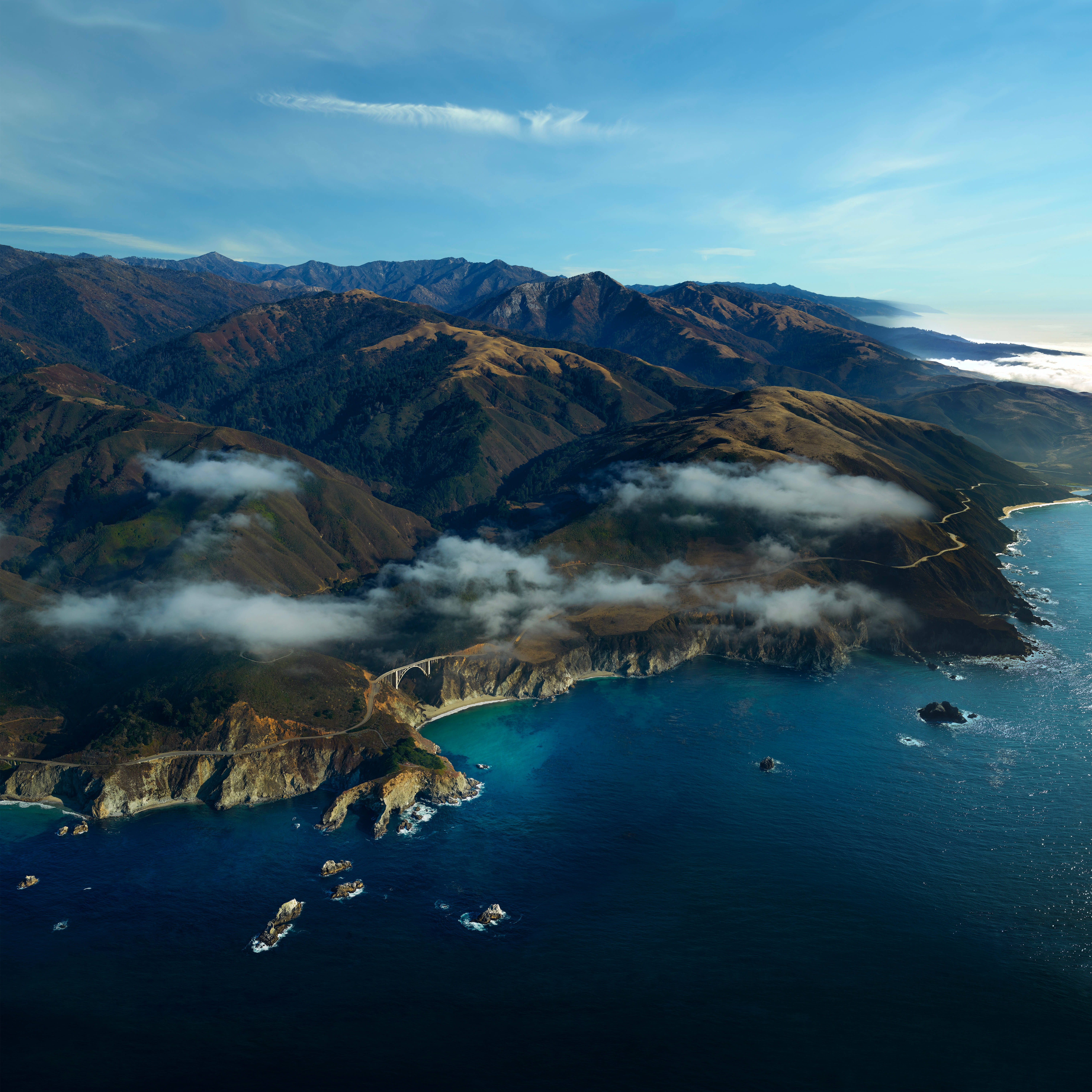 macOS Big Sur 山景 壁紙 — 鳥瞰圖：一條蜿蜒的沿海公路緊貼崎嶇懸崖，下方是碧綠海洋和起伏山丘，上方是清澈藍天。