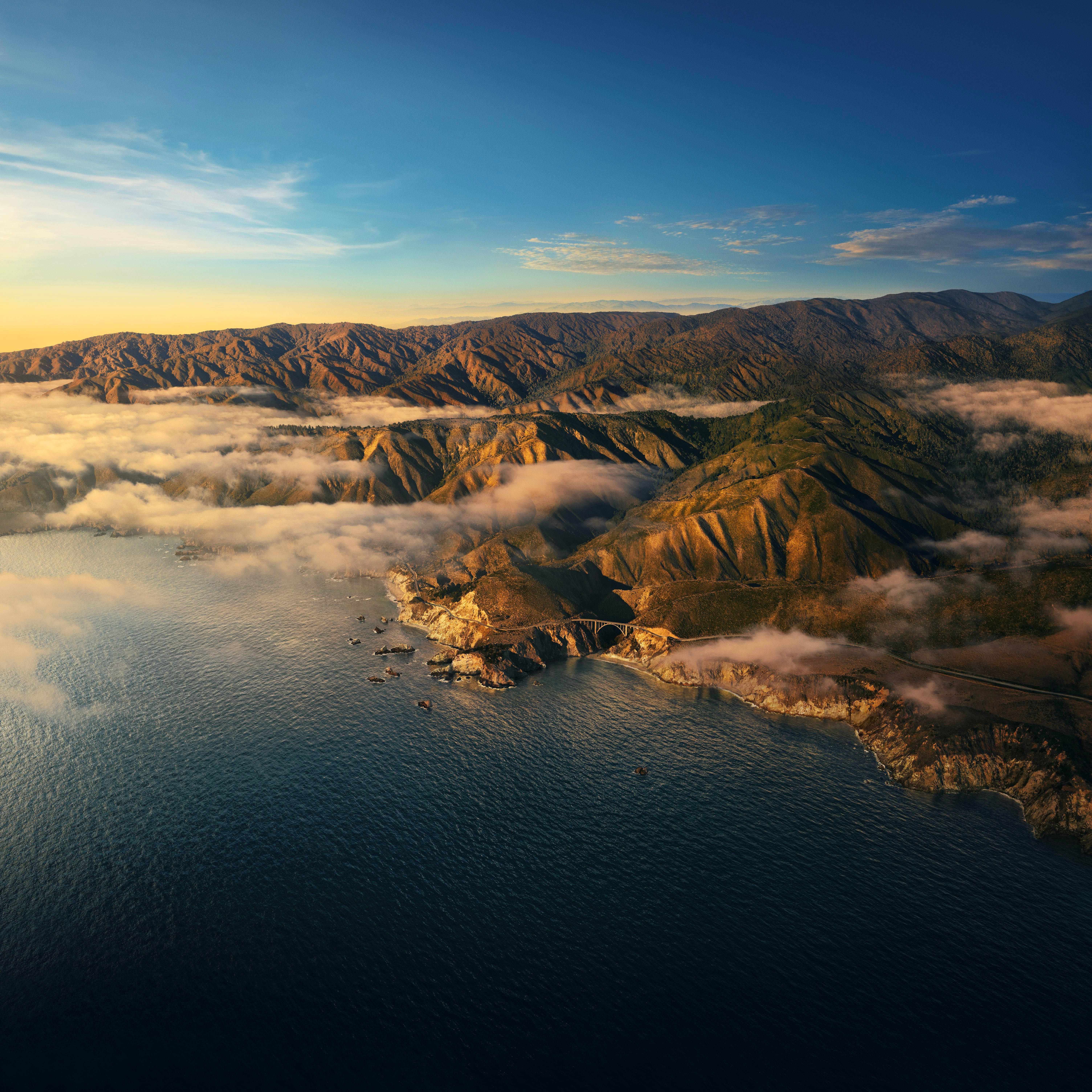 macOS Big Sur 山海日出 壁紙 — 崎嶇海岸山脈的鳥瞰圖，沐浴在金色日出光芒中，雲朵在下方飄移，深藍色海洋延伸至地平線。