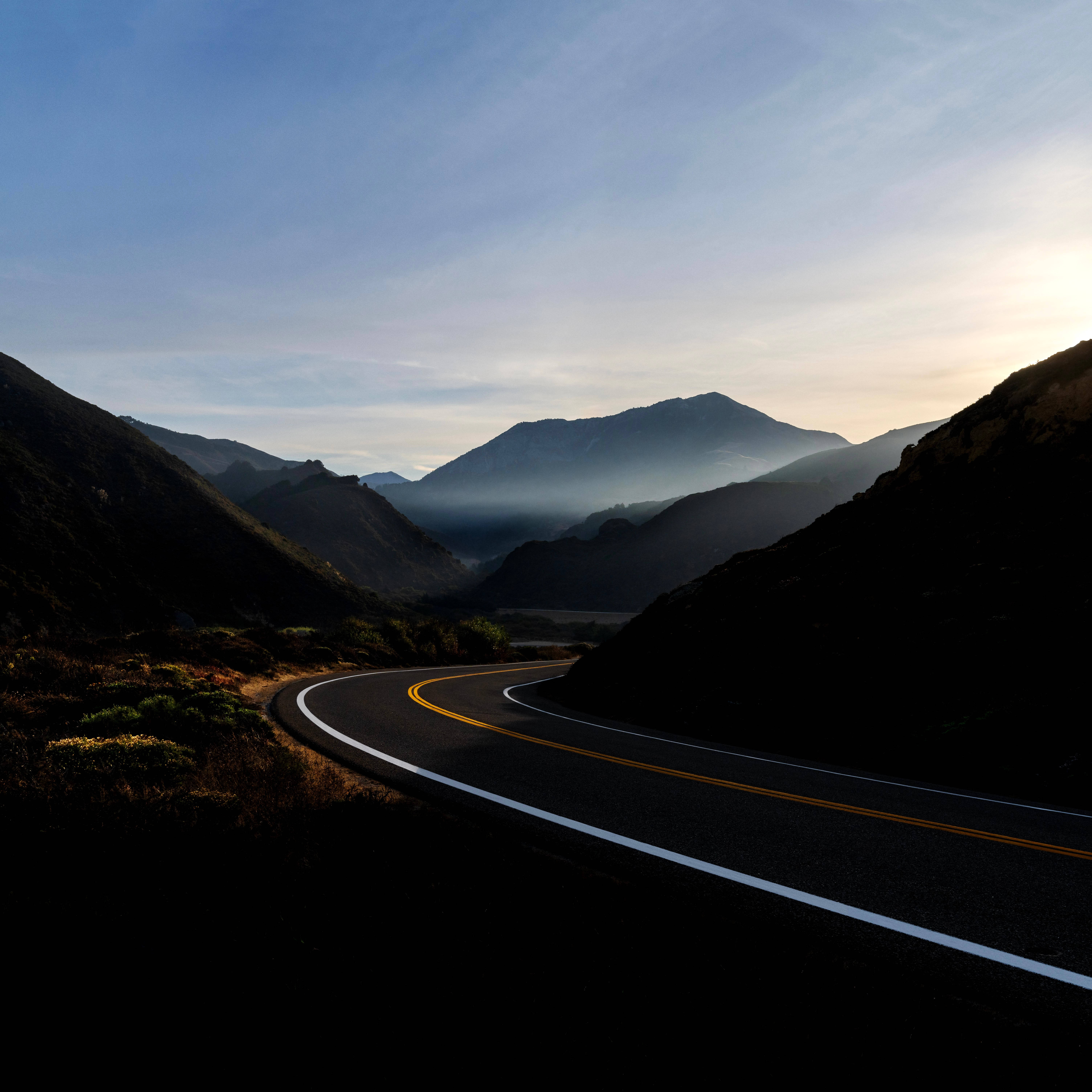 macOS Big Sur 山路 壁紙 — 一條蜿蜒的山路在柔和黎明天空下穿過陰影籠罩的山峰，下方是霧氣瀰漫的山谷。