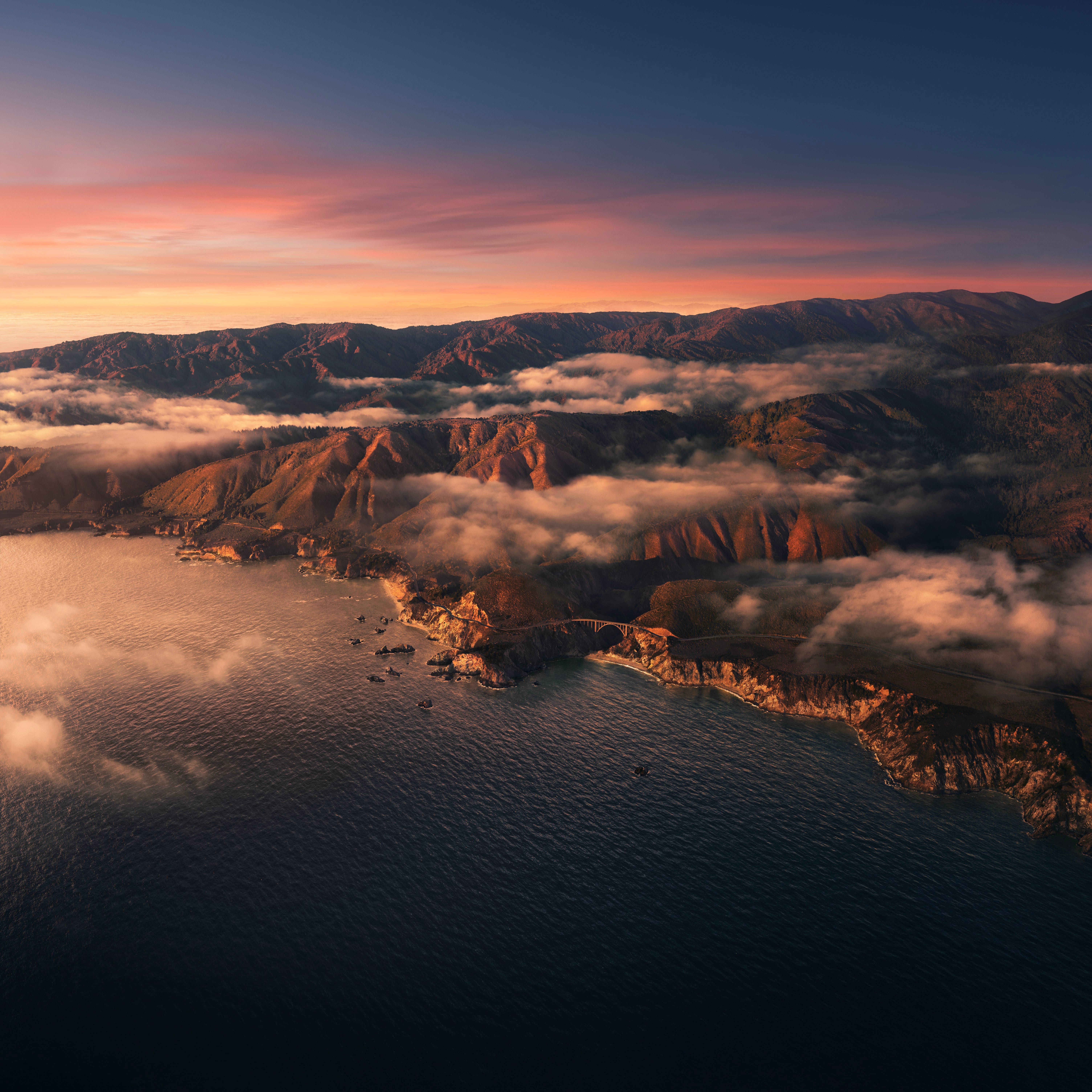 macOS Big Sur 日落海景 壁紙 — 日落時分海岸橋樑的鳥瞰圖，雲朵 nestled 在山脈與海洋之間。