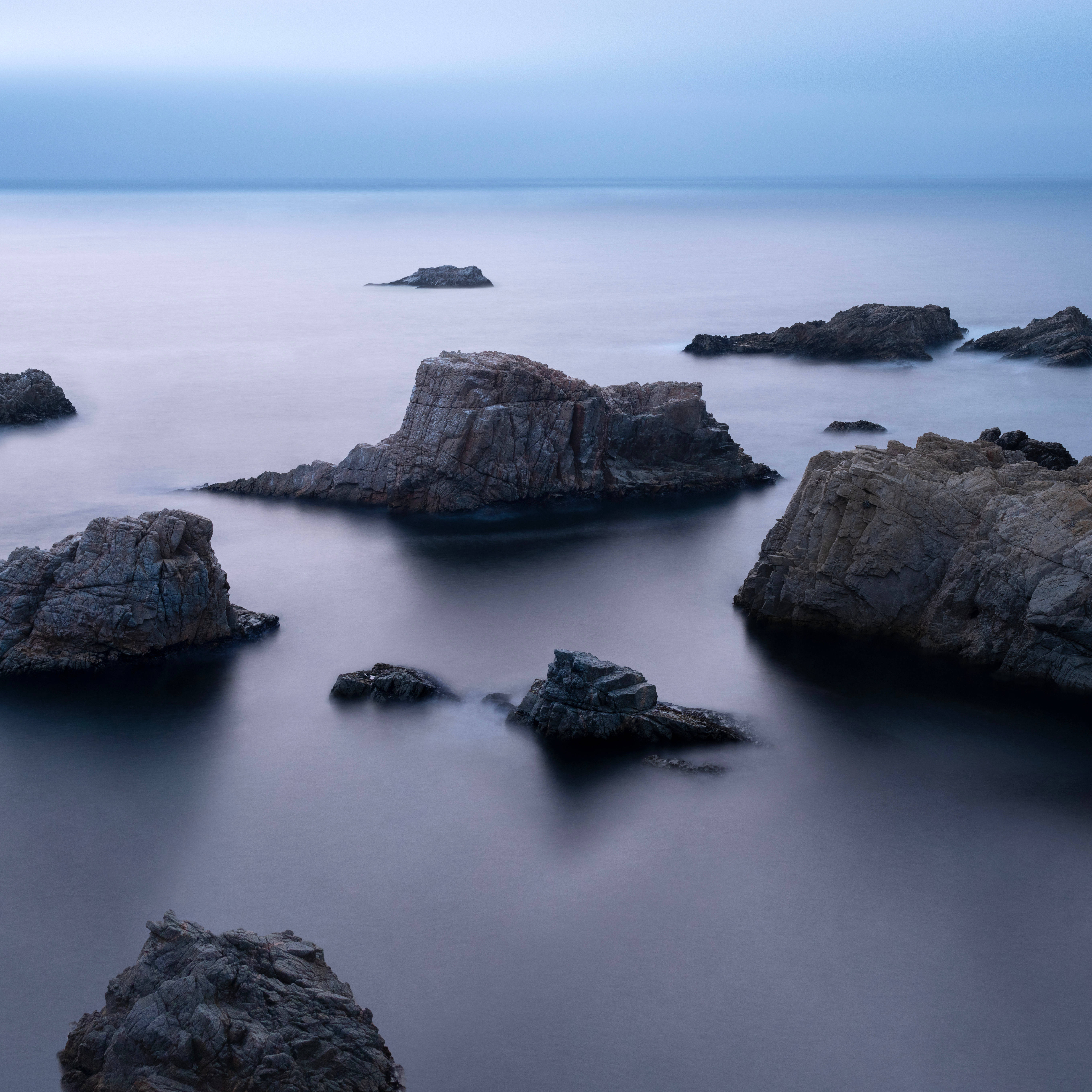 macOS Big Sur 海景 壁紙 — 寧靜的海岸岩石從平滑的藍色海水中浮現，在朦朧的暮色天空下。