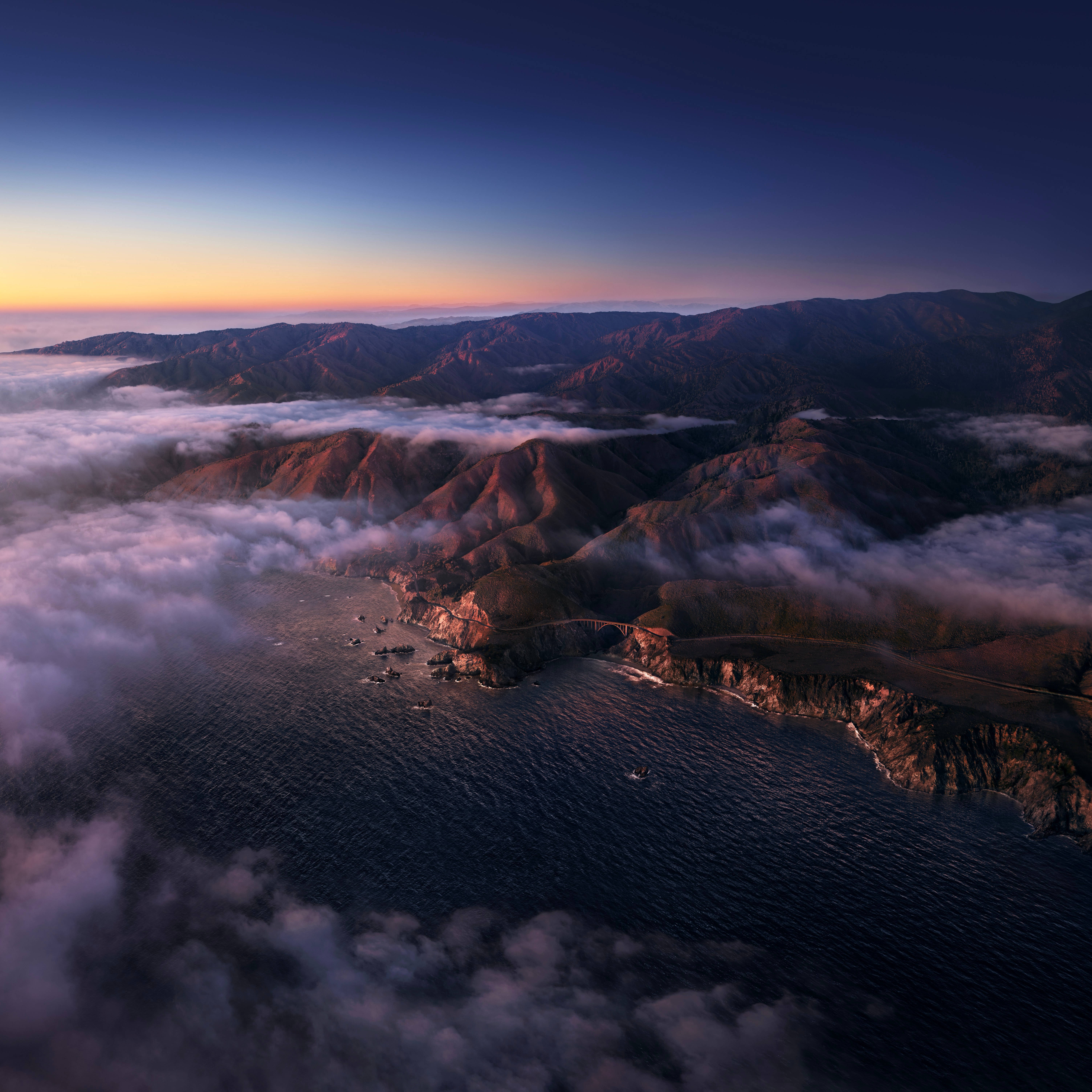 macOS Big Sur 黎明時分 壁紙 — 黎明時分海岸橋的鳥瞰圖，山脈在海霧和發光的天空上方升起。