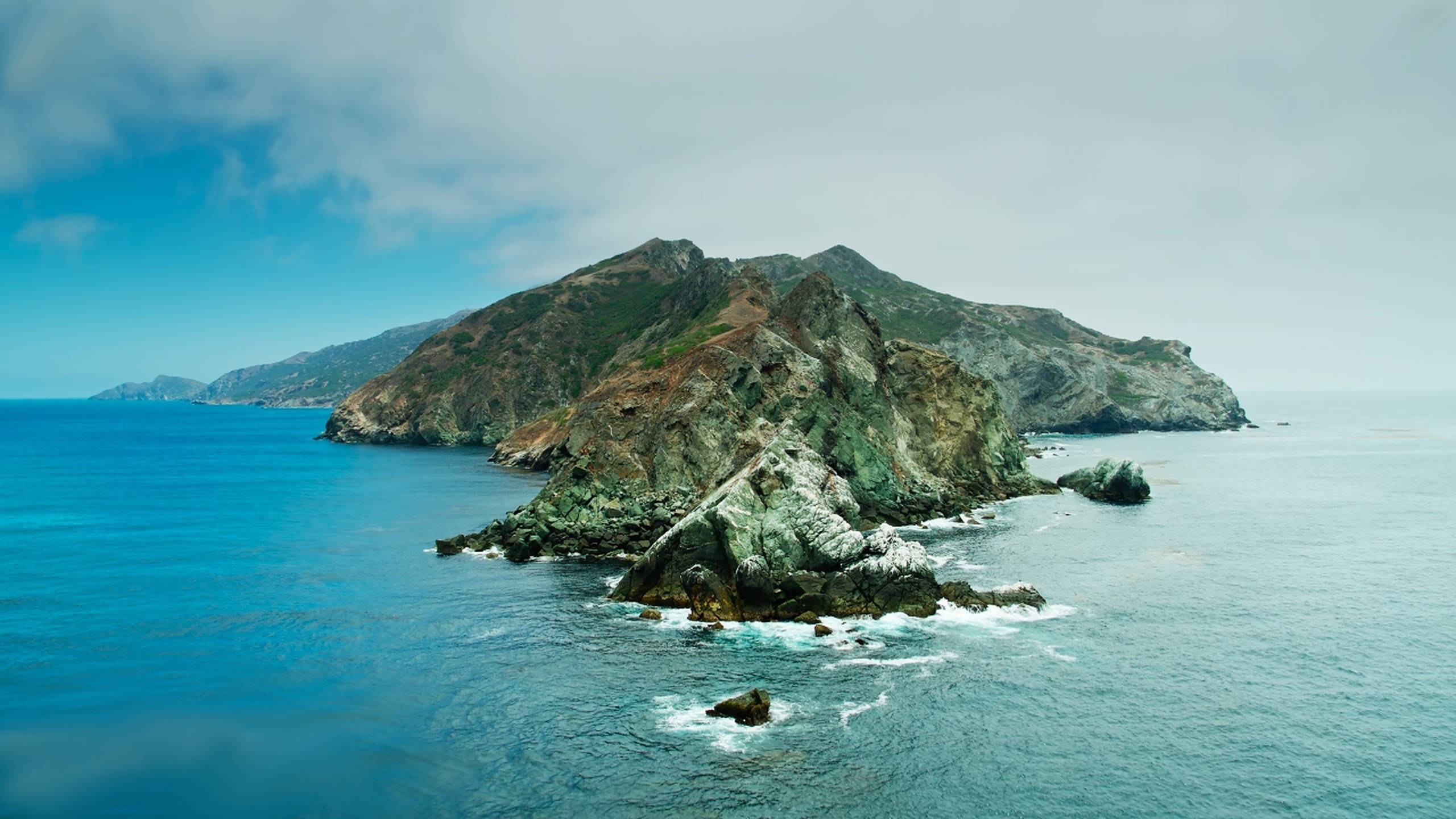 macOS Catalina 岩石山脈 壁紙 — 一座崎嶇的岩石島嶼從碧綠色的海水中升起，天空部分多雲，溫和的海浪拍打著它的底部。