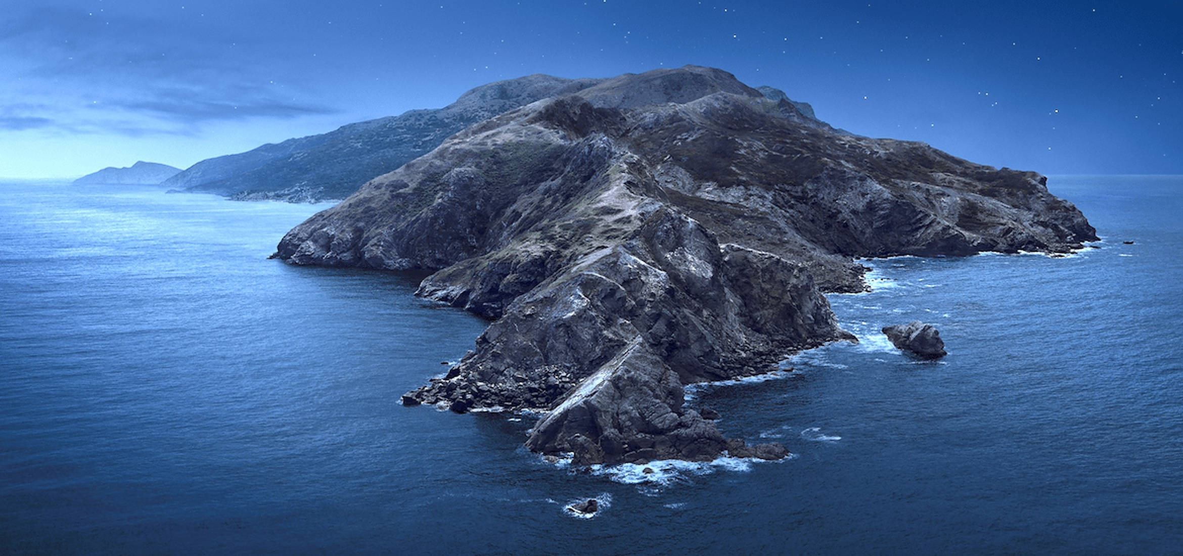 macOS Catalina 空中俯瞰圖 壁紙 — 一個崎嶇的岩石島嶼從深藍色的海洋中升起，在繁星點點的夜空下，地平線上有遙遠的山脈。