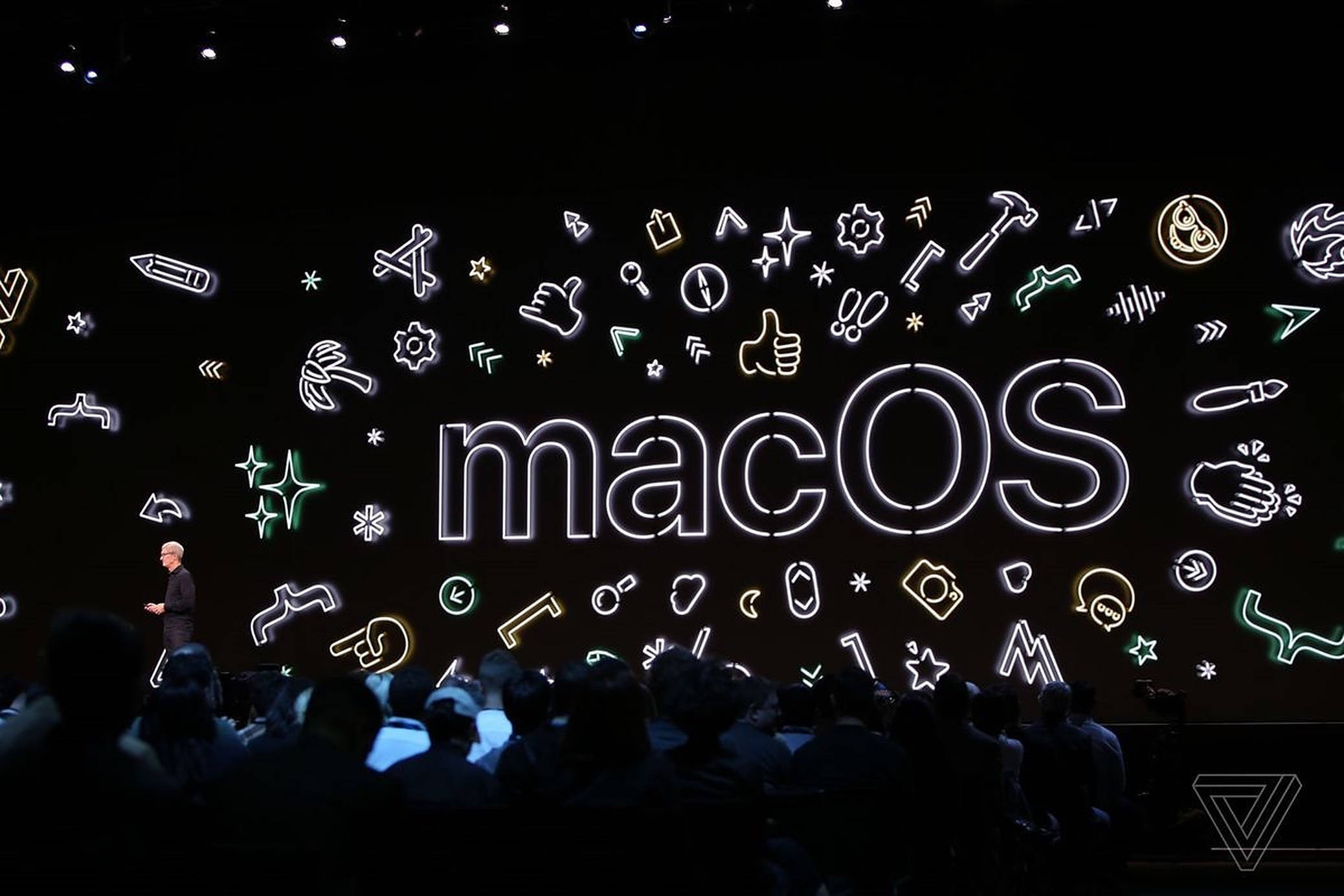 macOS Catalina 舞台現場 壁紙 — 一位演講者在黑暗的舞台上，在觀眾面前展示一個被科技圖標環繞的發光霓虹macOS標誌。