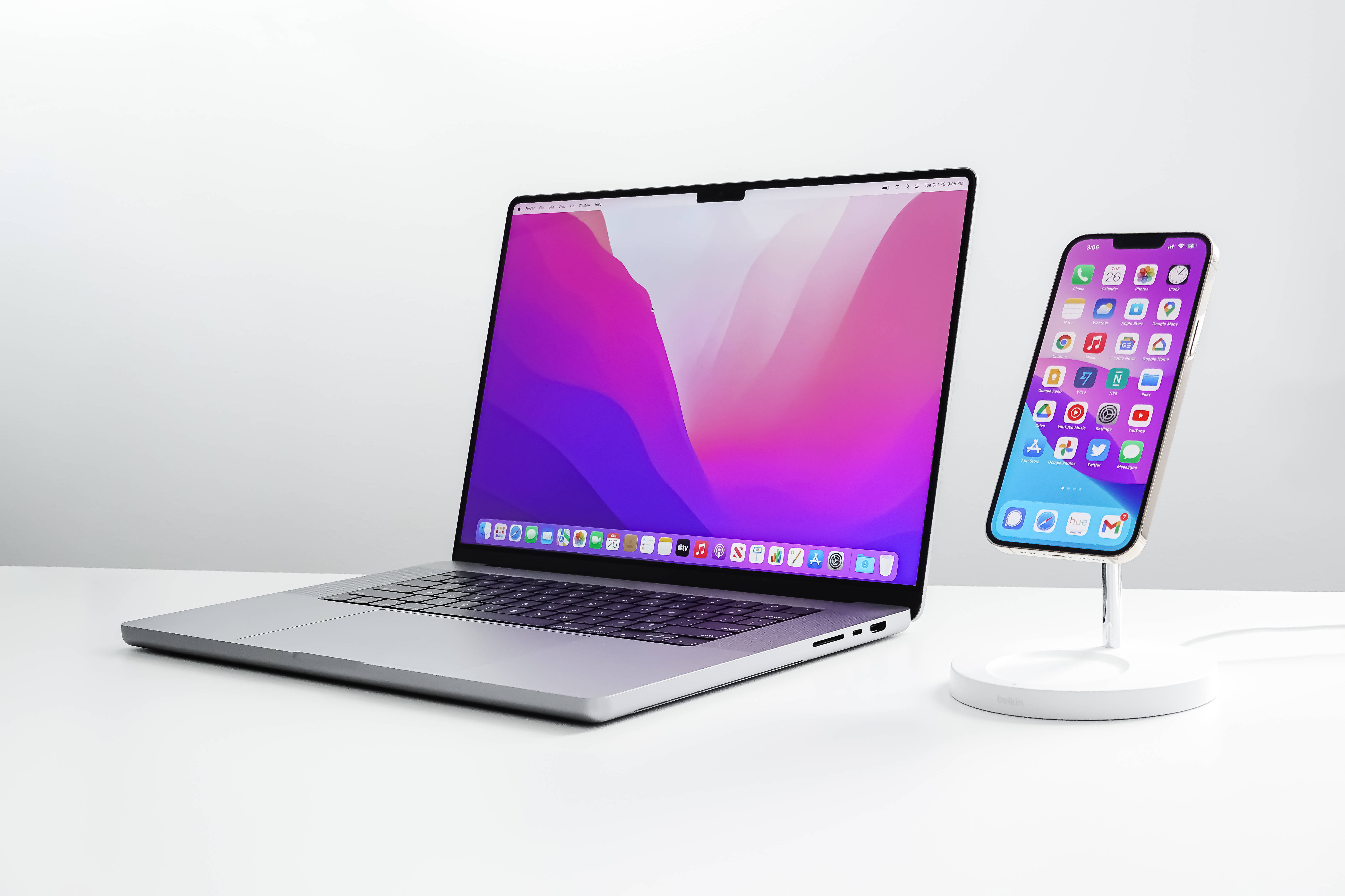 macOS Monterey 在 Apple 設備上 壁紙 — 一部時尚的銀色手提電腦和iPhone，帶有鮮豔的紫粉漸變屏幕，並排放置在簡約的白色桌子上。