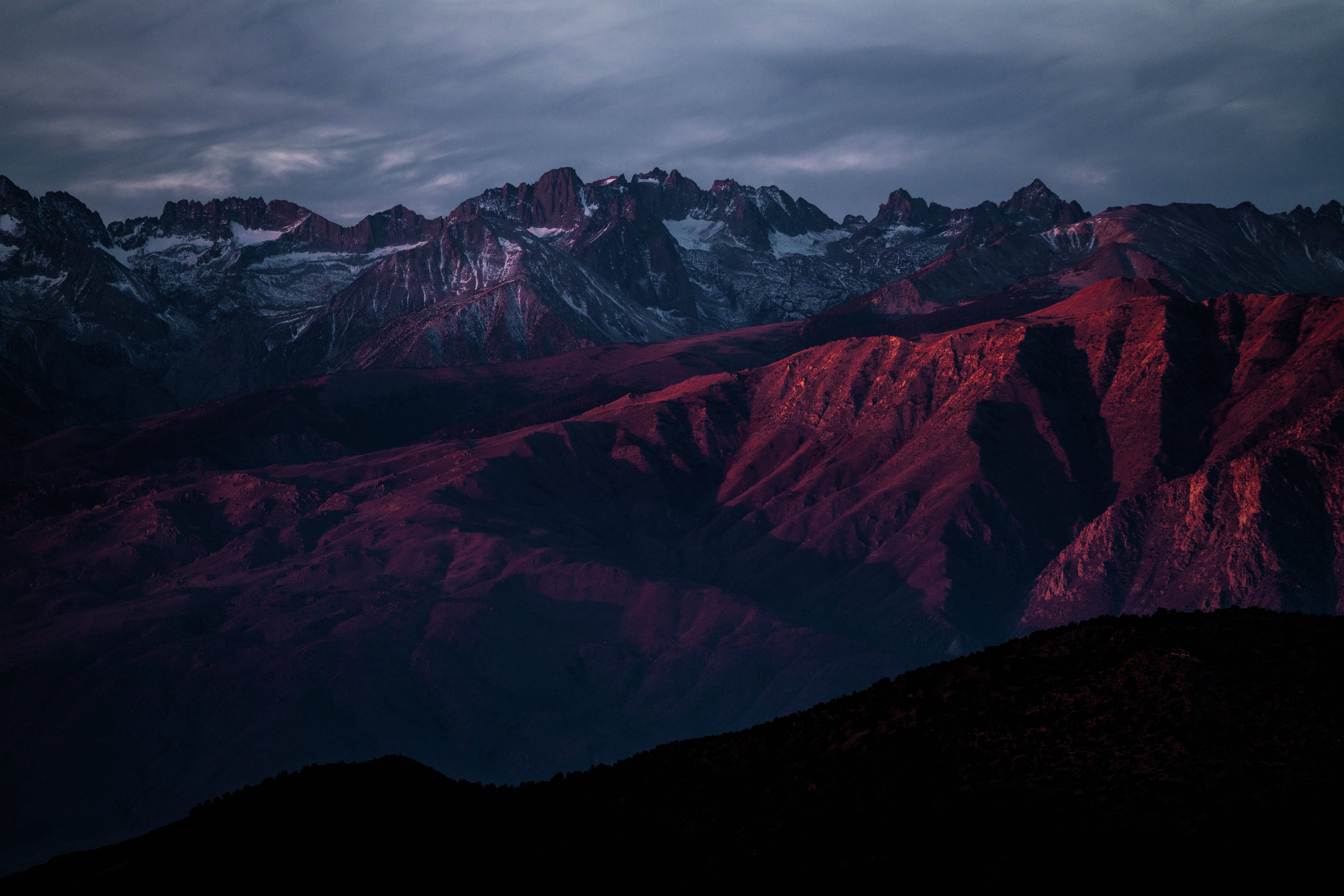macOS 預設壁紙：壯麗山景 壁紙 — 一條戲劇性的山脈沐浴在深紅色的暮光中，白雪覆蓋的山峰在陰沉的灰色天空下。