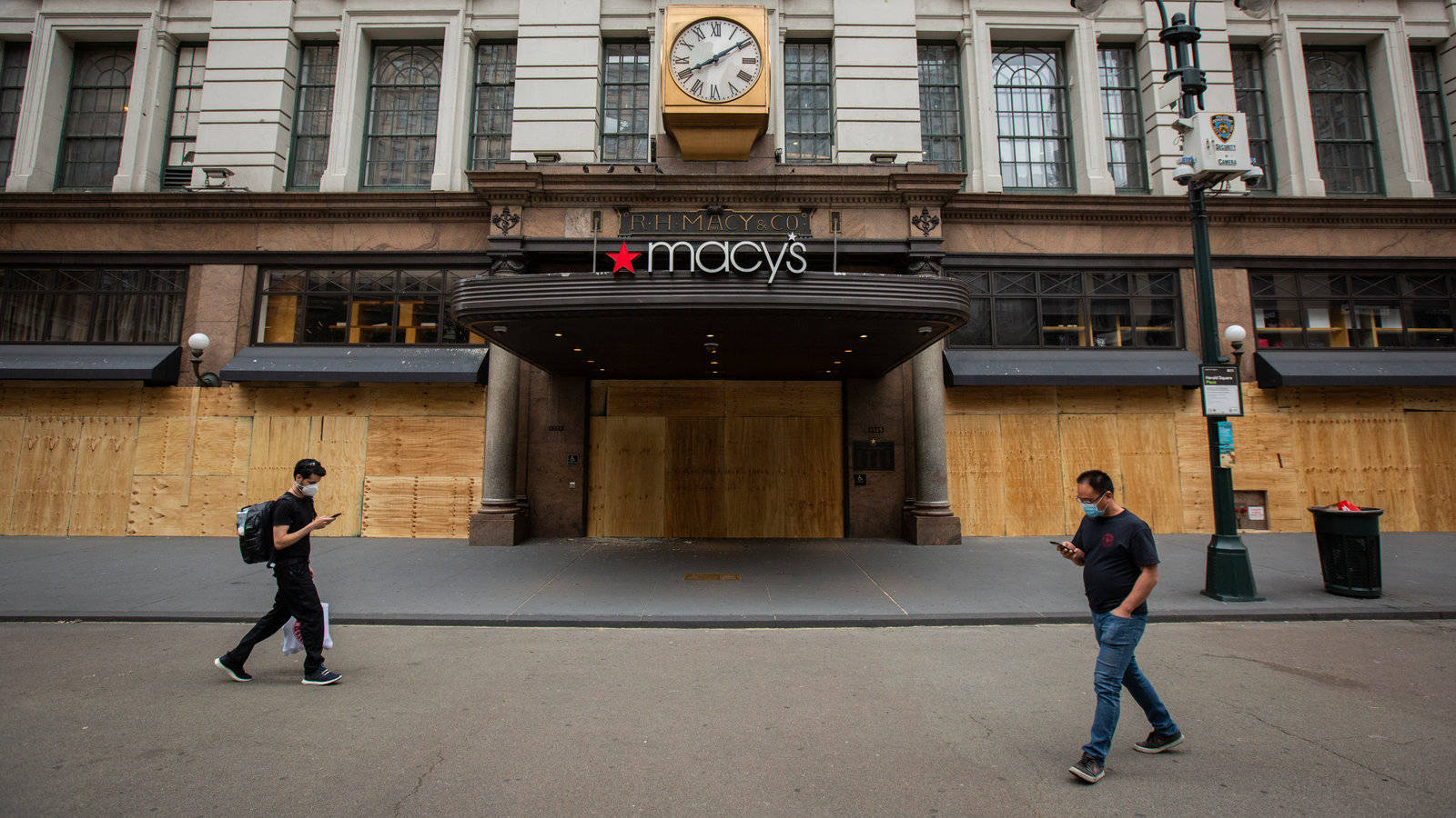 兩名戴著口罩的人走過一間用木板封住的Macy's商店，上方有時鐘。