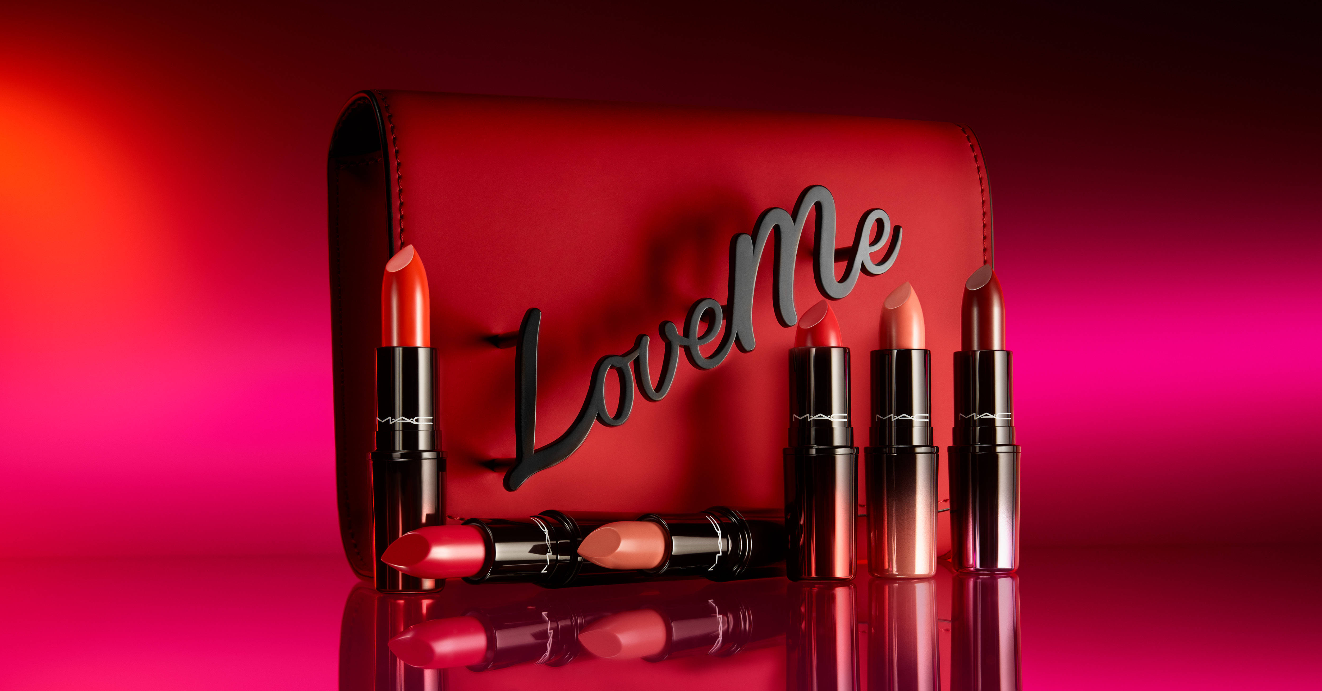 MAC彩妝Love Me系列 壁紙 — 一個時尚的紅色MAC唇膏盒，上面有黑色的「LoveMe」字樣，周圍是鮮豔的紅色和粉紅色唇膏，放在漸變反射表面上。