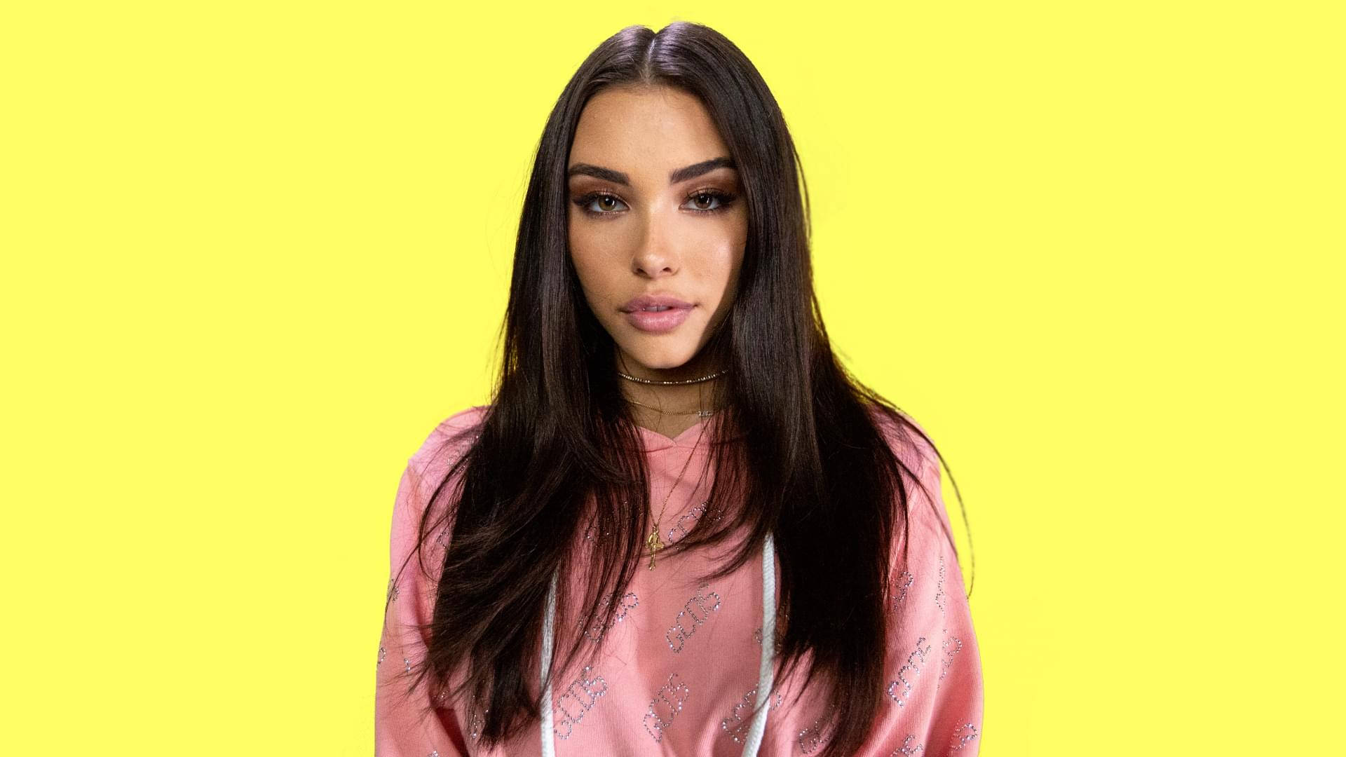 Madison Beer 黃色背景 壁紙 — 一位長黑髮的年輕女性穿著粉紅色連帽衫，背景是明亮的黃色。