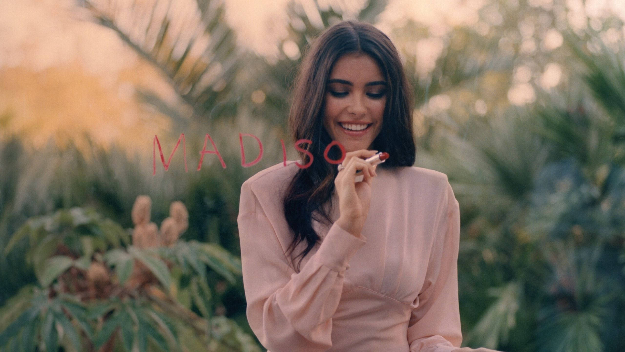 年輕 Madison Beer 壁紙 — 一位微笑的女士身穿粉紅色襯衫，在柔焦的熱帶花園背景下拿著一支香煙。