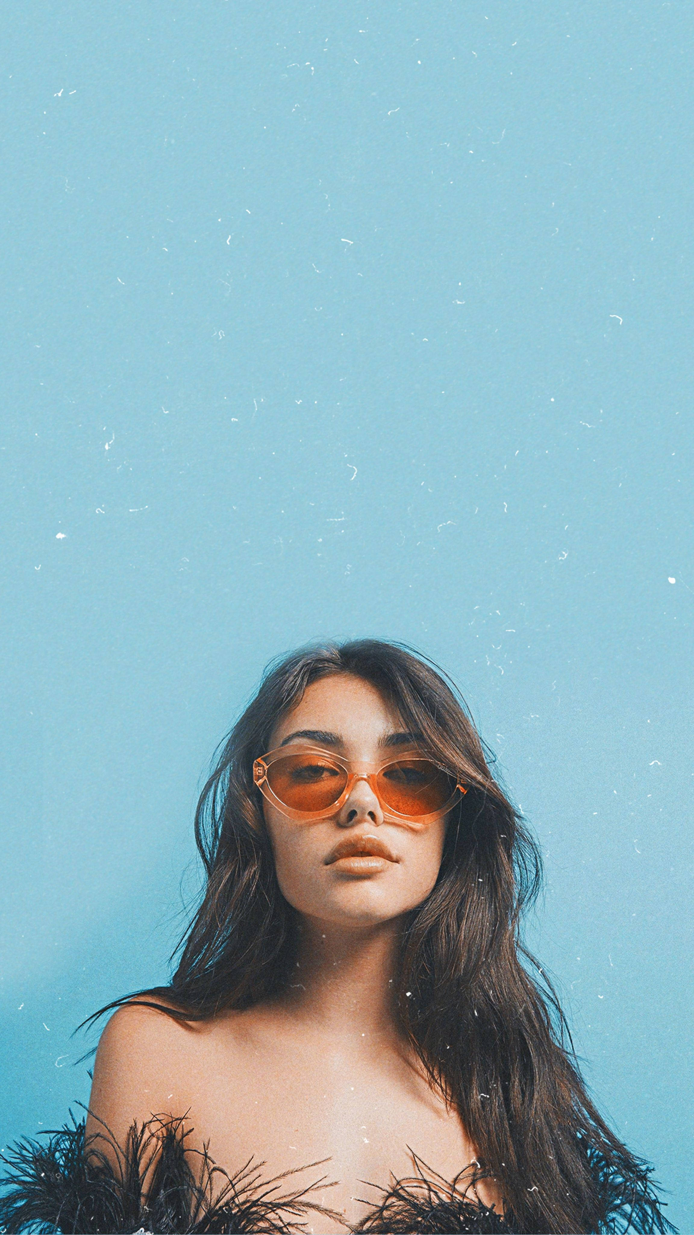 Madison Beer 藍色美學風 壁紙 — 一位長黑髮的女性戴著橙色太陽眼鏡，背景是帶紋理的淺藍色。