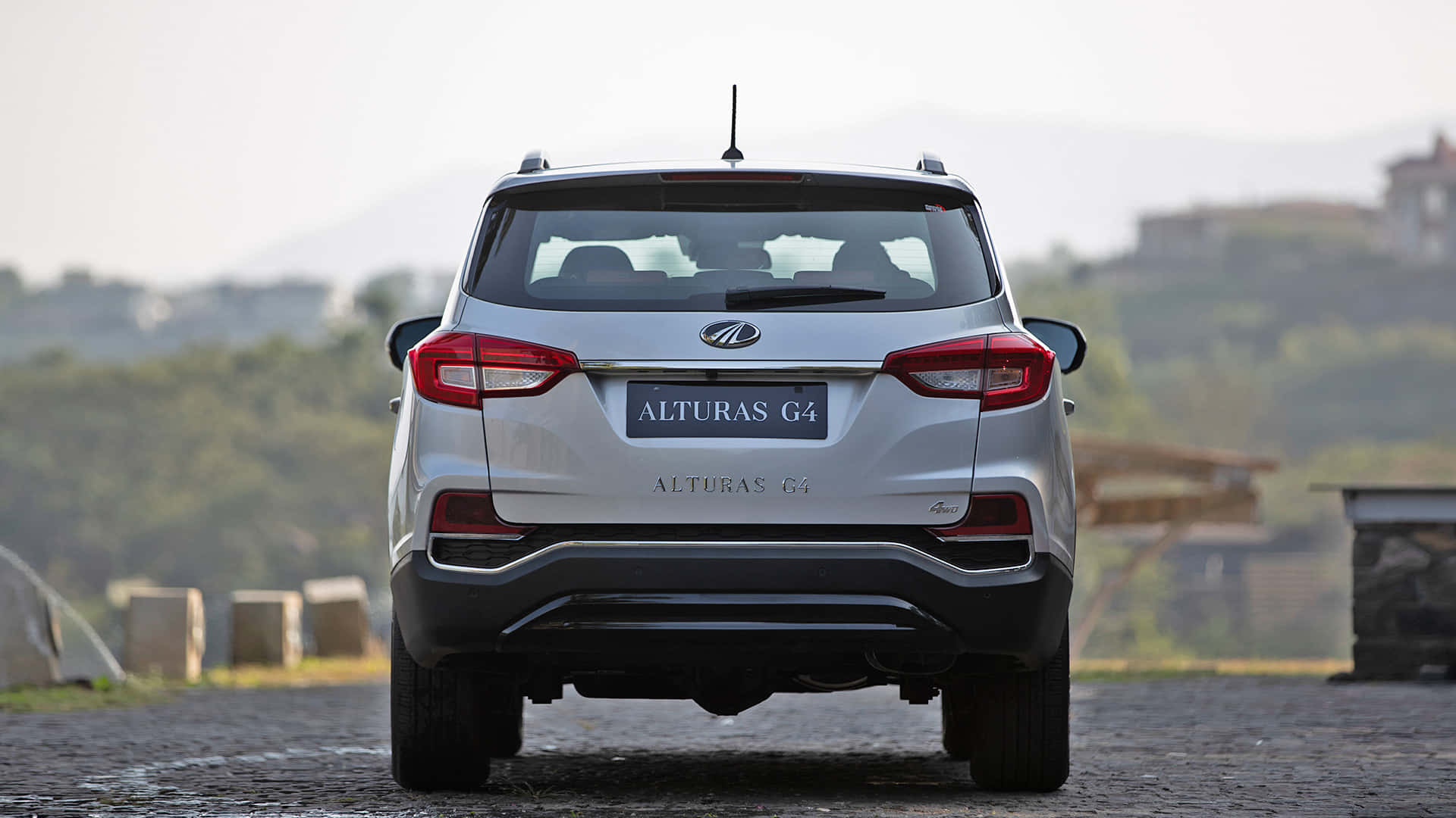 Mahindra Alturas G4車尾視角 壁紙 — 從後方觀看銀色 Alturas G4 SUV 停泊在鵝卵石路上，背景有朦朧的山丘和建築物。
