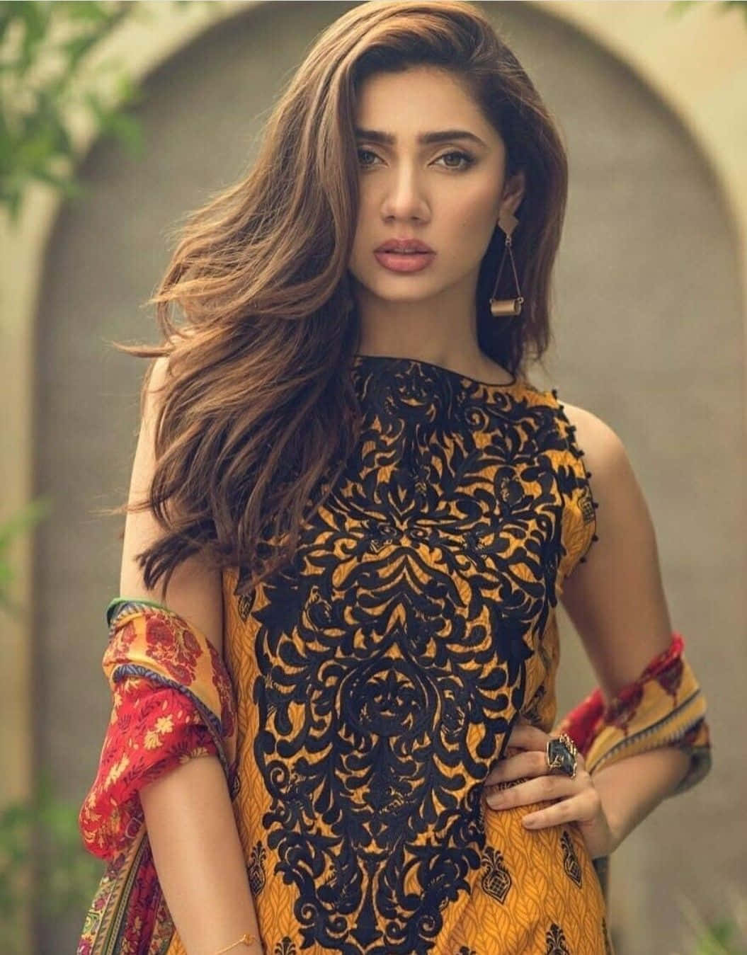 巴基斯坦女孩Mahira Khan 黃黑禮服照片 壁紙 — 一位擁有長波浪棕色頭髮的女性，穿著鮮豔的黃黑圖案連衣裙，搭配紅色花卉袖子，背景柔和呈拱形。