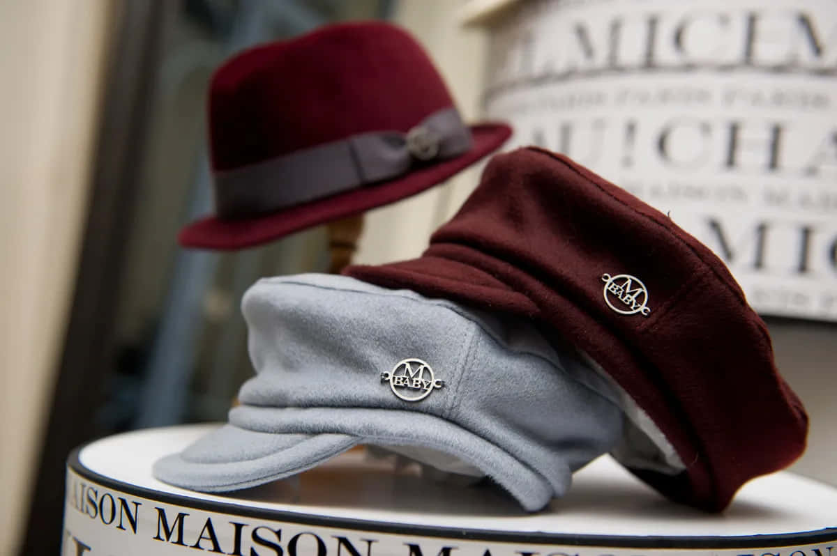 Maison Michel 帽款展示 壁紙 — 三頂時尚的酒紅色和灰色帽子放在品牌展示架上，背景柔和。