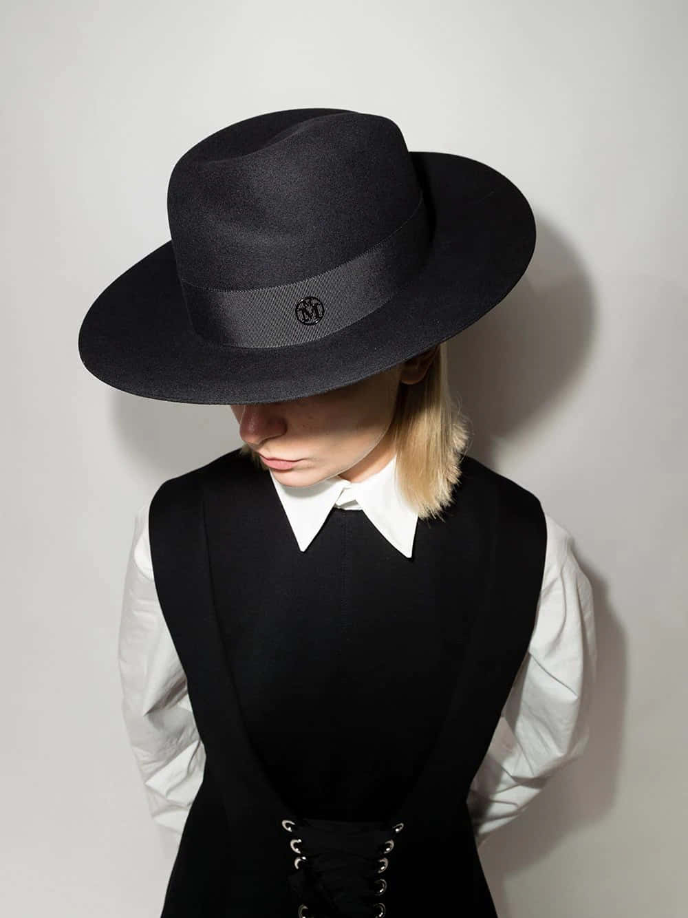 Maison Michel 黑色毛呢 Fedora 帽 壁紙 — 一個人戴著黑色軟呢帽、穿著背心和白色襯衫，靠著素牆站立，臉部部分被帽子遮擋。