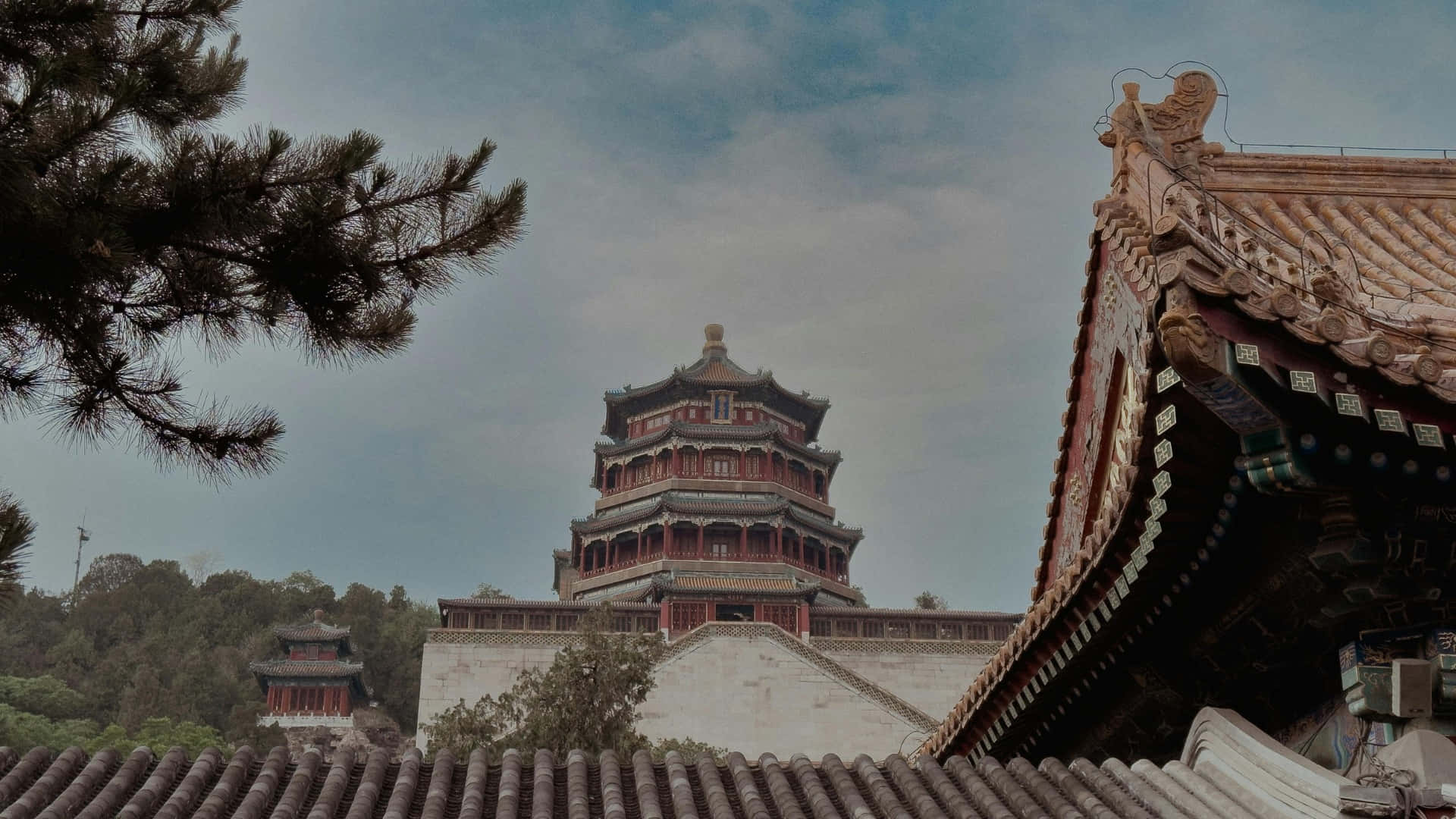  壁紙 — 一座雄偉的傳統中式寶塔在雲天下聳立於瓦頂和松樹之上。